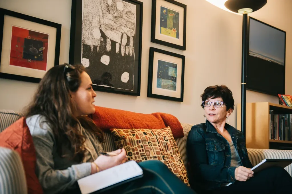 Two women sitting on a sofa having a conversation, one is holding a notebook and pen, and the other is holding a tablet, with framed artwork on the wall behind them.