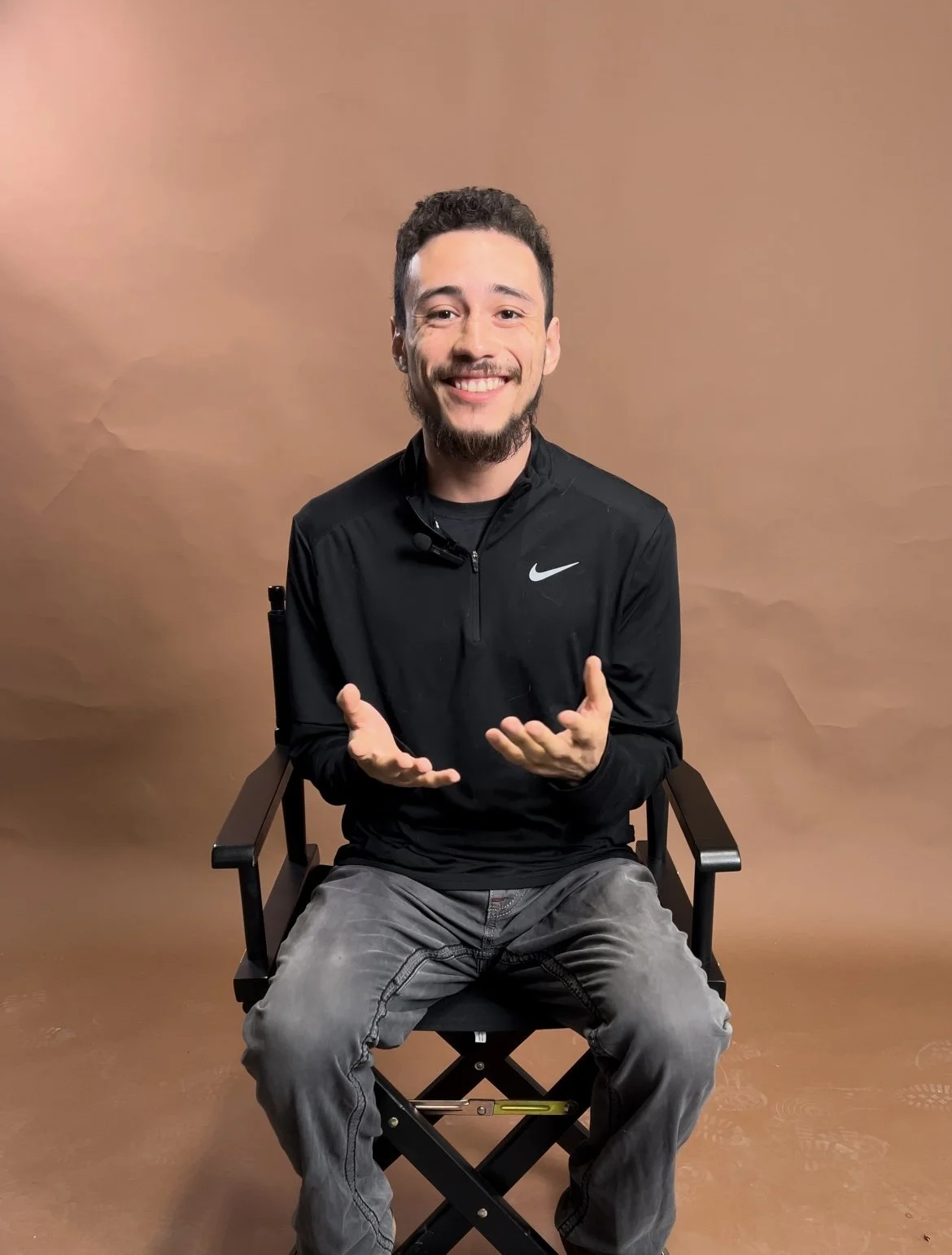 A man with short dark hair, a beard, and wearing a black Nike jacket, sitting on a director's chair, smiling and gesturing with his hands, against a brown background.