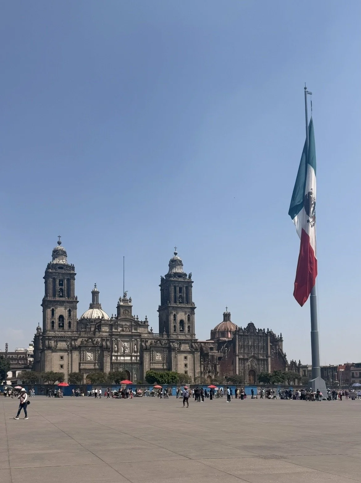 Large Mexican flag flying in front of the Mexico City Metropolitan Cathedral with people in the plaza.