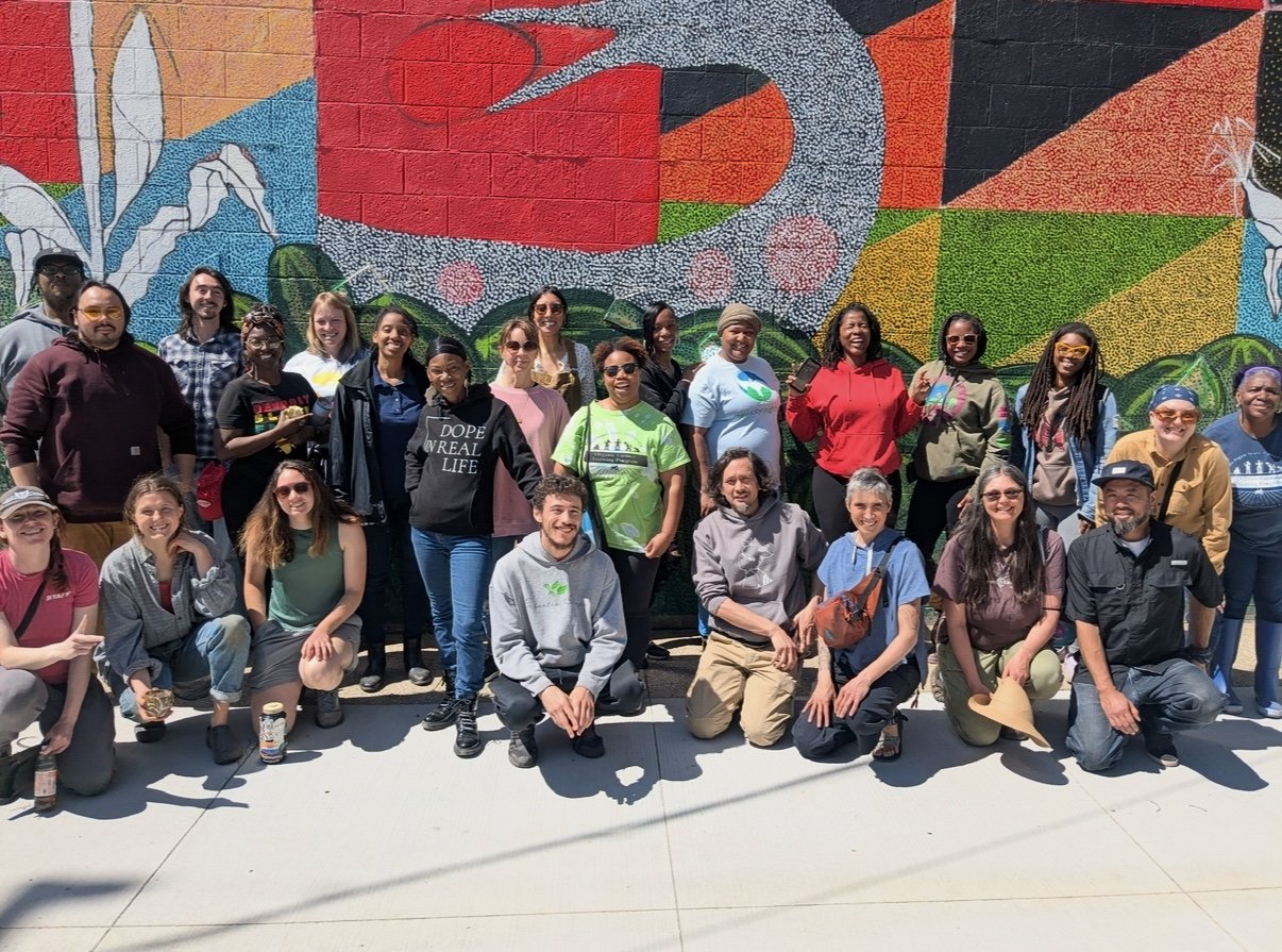Group of diverse people posing in front of a colorful mural on a brick wall. The mural features various bright colors and patterns.