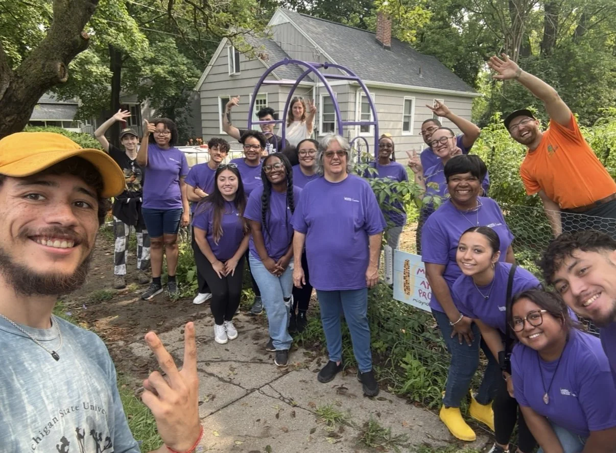 Group of people smiling outdoors in front of a house, some wearing purple shirts, with a garden and trees in the background.