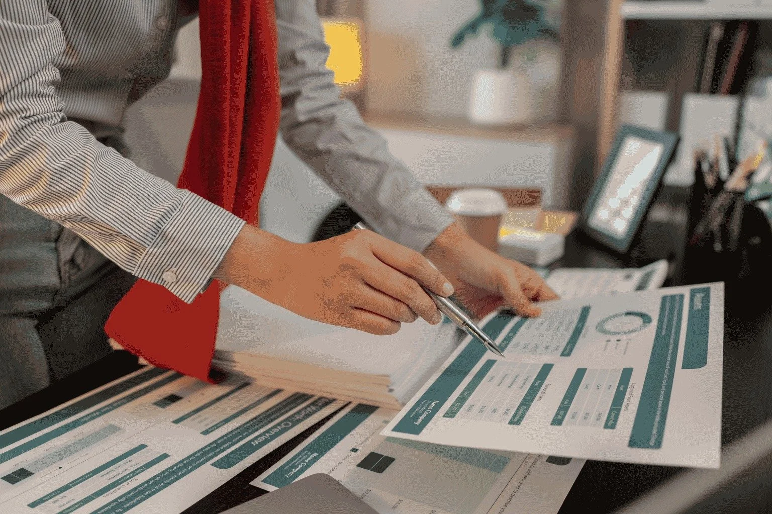 Close-up of a person in a gray striped shirt and red blazer holding a pen and filling out a form or worksheet on a desk, with papers and a tablet in the background.
