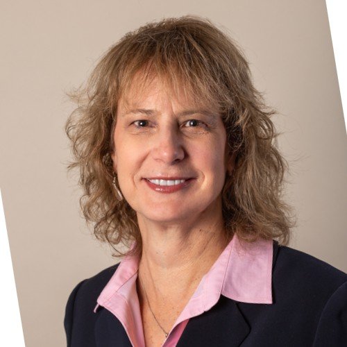 A woman with curly blonde hair, wearing a navy blazer and pink collared shirt, smiling against a light background.