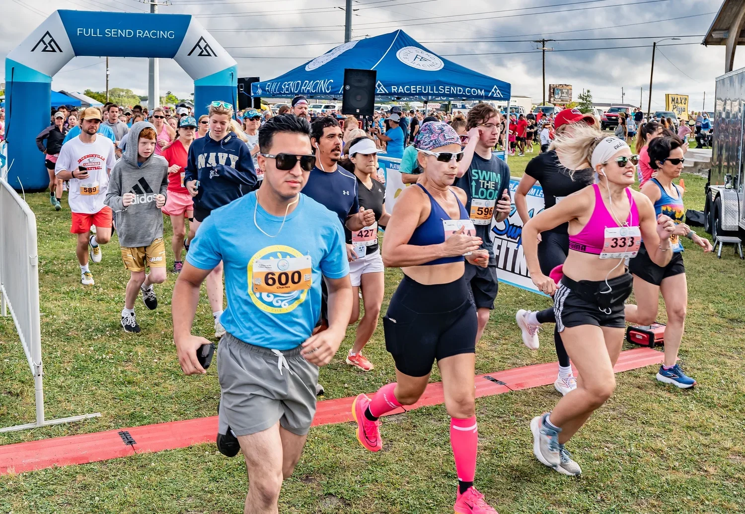 People participating in a running event, exiting the starting line, with a blue archway and tents in the background, on a grassy field under an overcast sky.