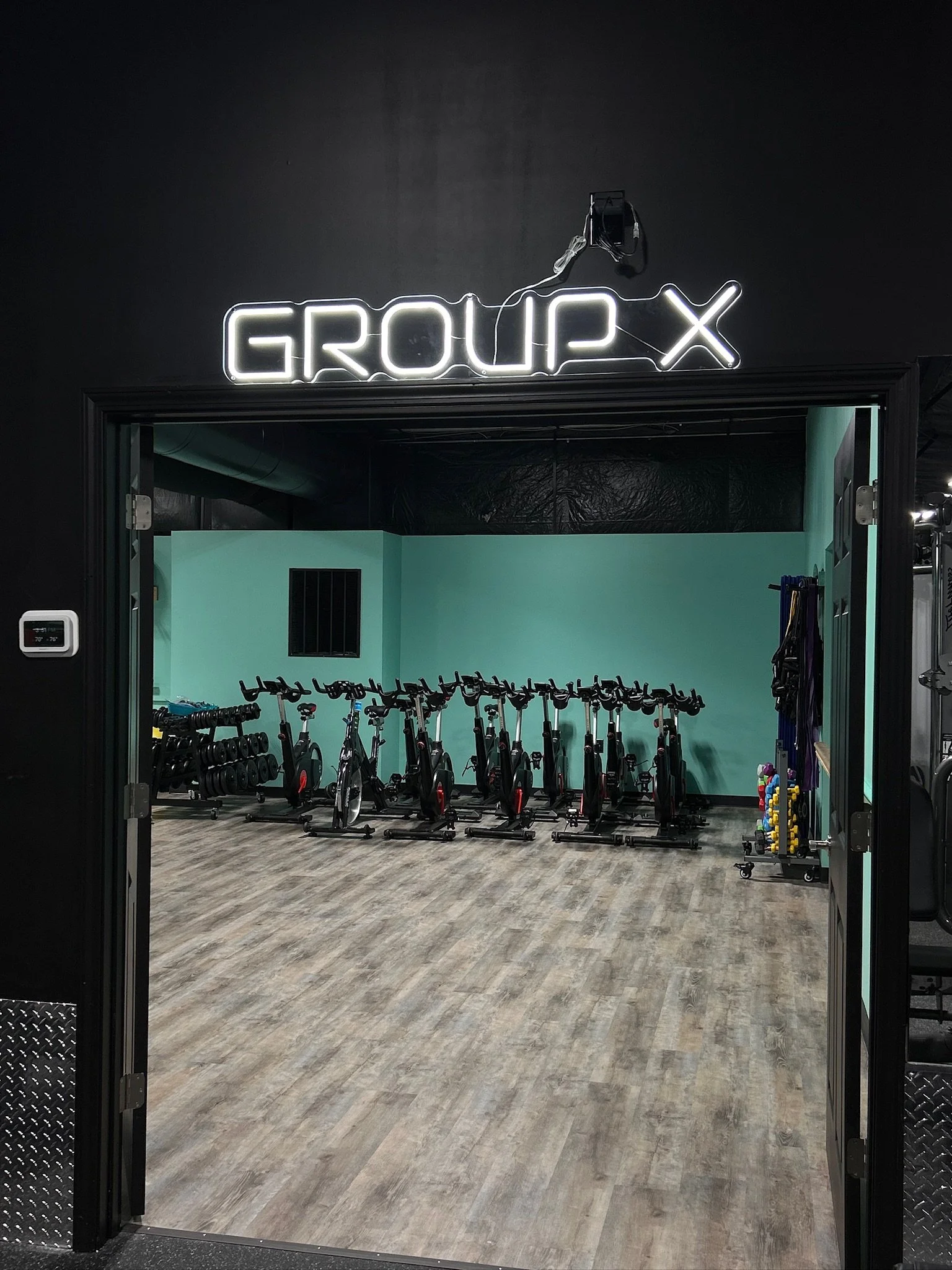 View of a fitness studio with multiple stationary bikes lined up, visible through an open door with a neon sign that says 'GROUP X' above the entrance.