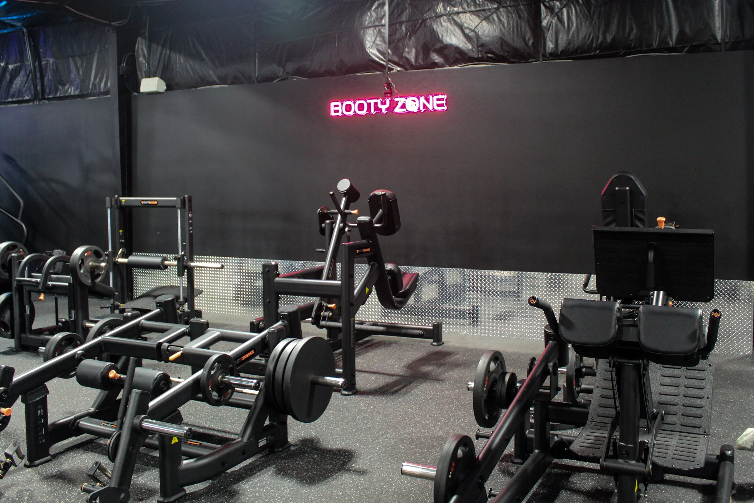 A gym with fitness equipment such as weights, treadmills, and exercise machines, and a neon sign on the wall that reads 'Booty Zone' in pink.