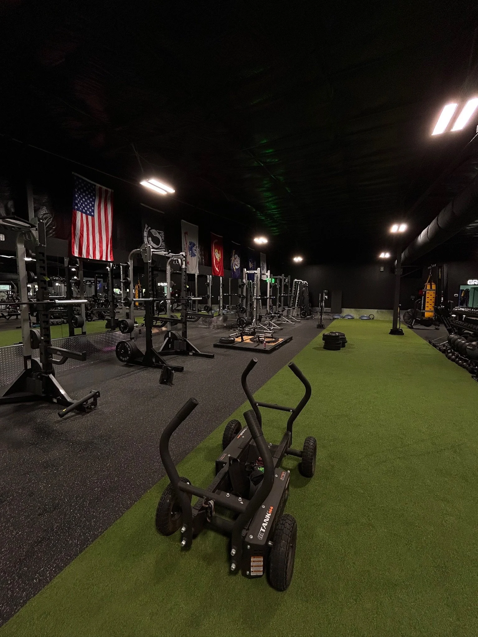 A gym with weightlifting equipment, American flags, and banners on the wall. There is a green artificial turf on the floor and black rubber flooring. Fitness equipment like a sled, barbells, and weight plates are visible.