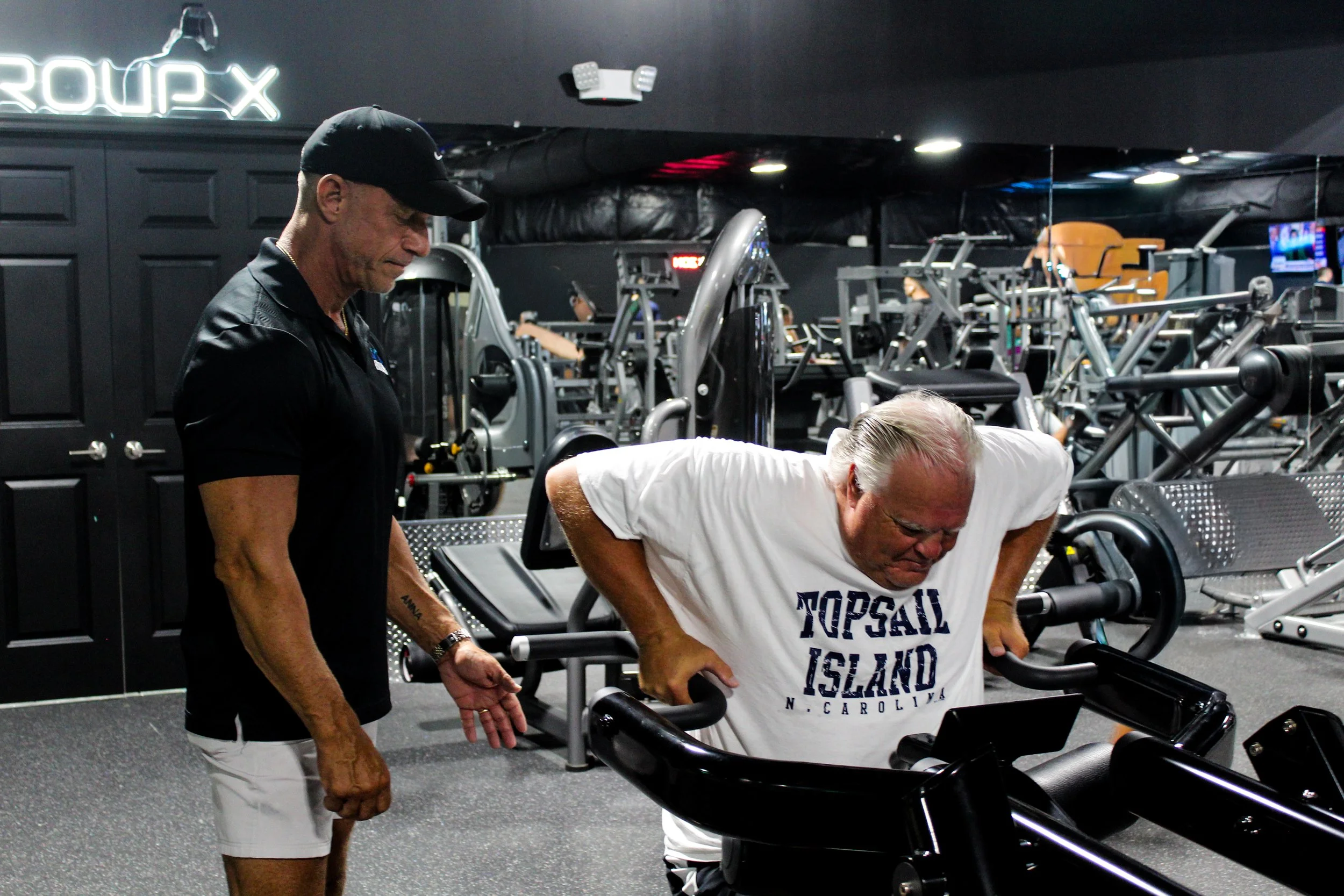 Two men working out at a gym, with one man in a white T-shirt leaning forward on exercise equipment and the other man in a black cap and shirt standing nearby.