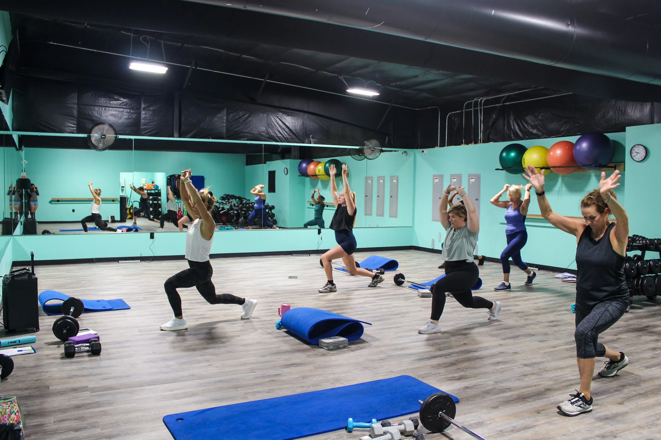 Group of people participating in a fitness class in a gym, performing exercises with their arms raised, surrounded by workout equipment like mats, weights, and balls.