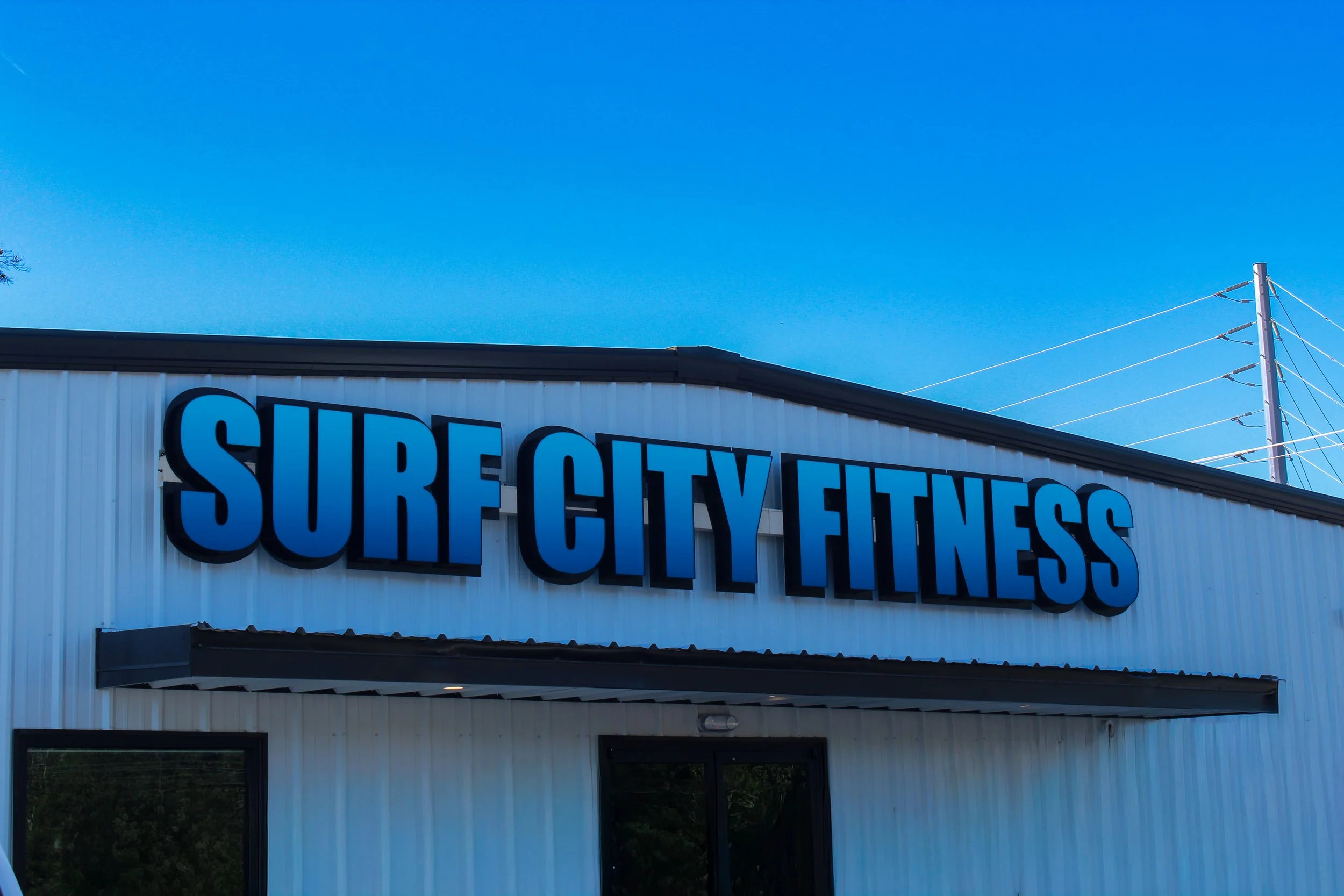 Storefront with a blue sign that reads "SURF CITY FITNESS" on a white building with a black roof and large windows, against a clear blue sky.