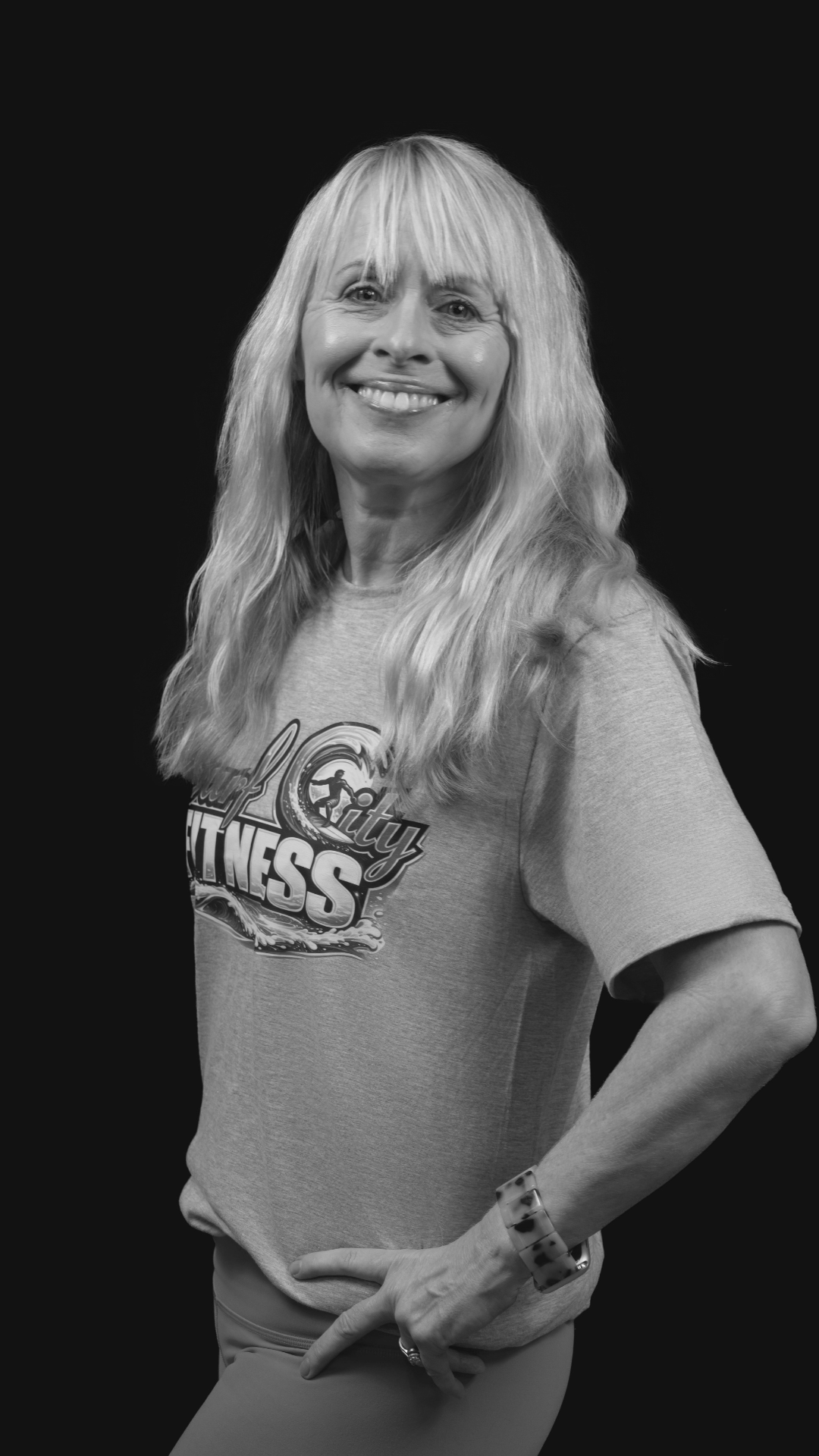 Black and white photo of a smiling woman with long, wavy hair, wearing a t-shirt with fitness-themed graphics, posing with one hand on her hip and the other resting on her thigh, against a dark background.