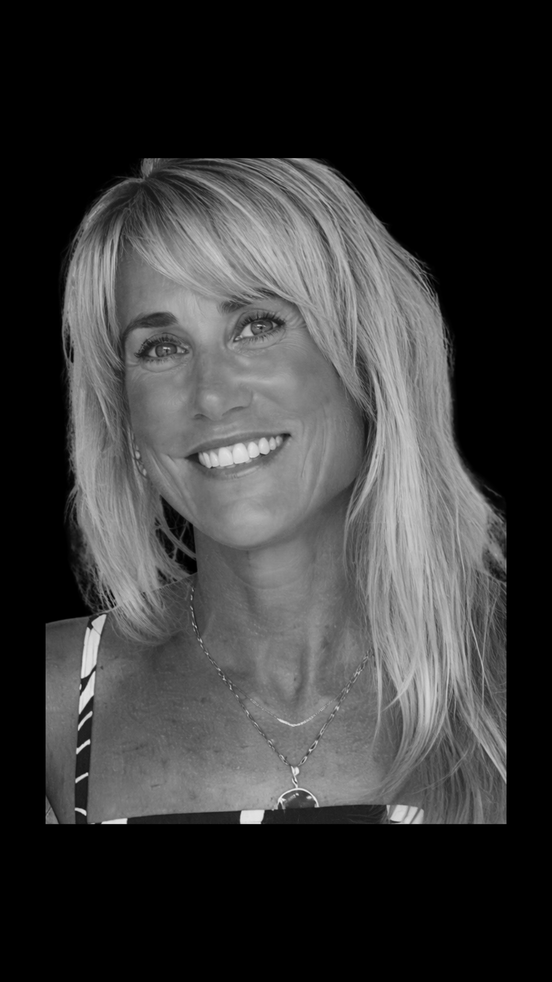 Black and white portrait of a smiling woman with shoulder-length hair, wearing a striped top, necklaces, and earrings, against a solid dark background.