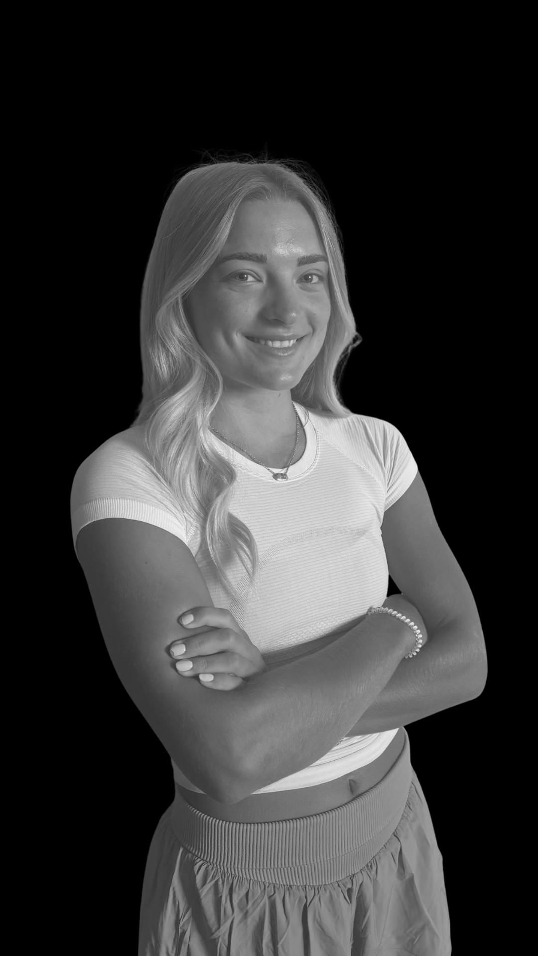 Black and white portrait of a young woman with long wavy hair, smiling, wearing a short-sleeved shirt, a necklace, a bracelet, and a skirt, crossing her arms, against a black background.