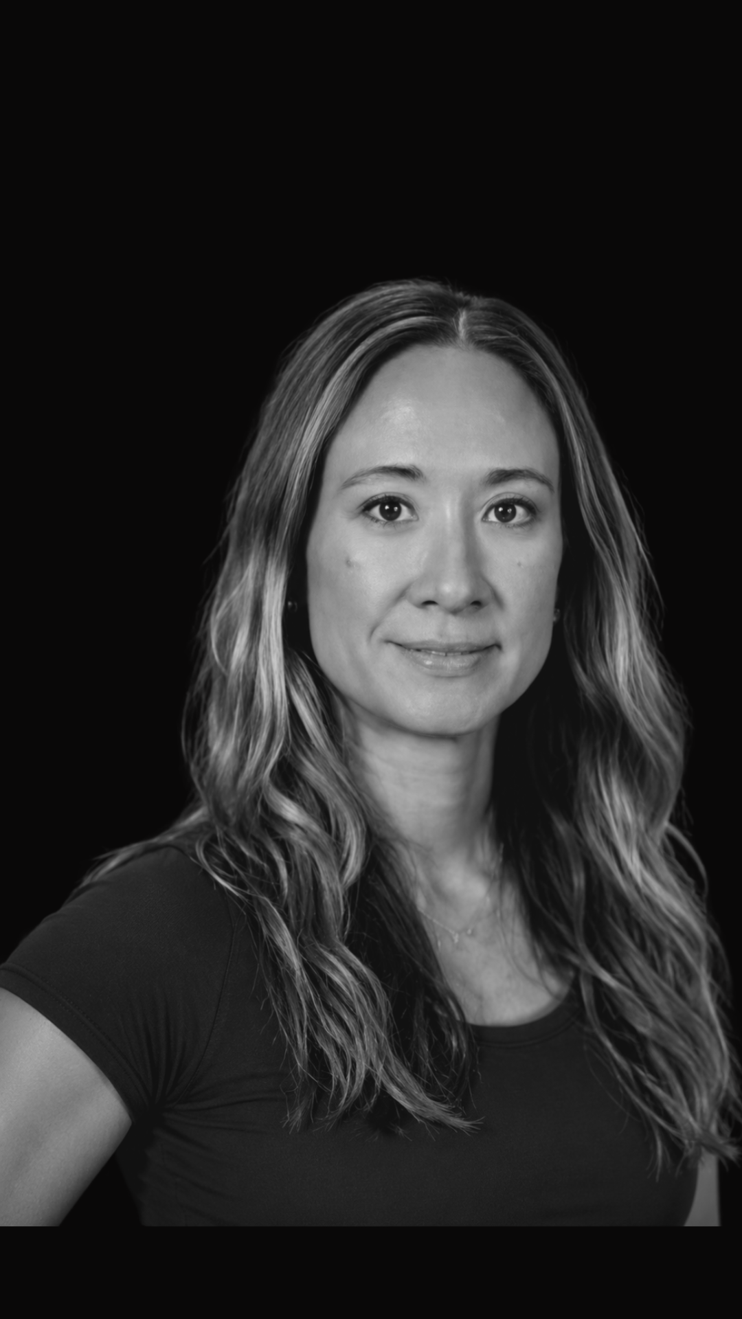 Black and white portrait of a woman with long wavy hair, wearing a black shirt, smiling slightly, against a plain dark background.