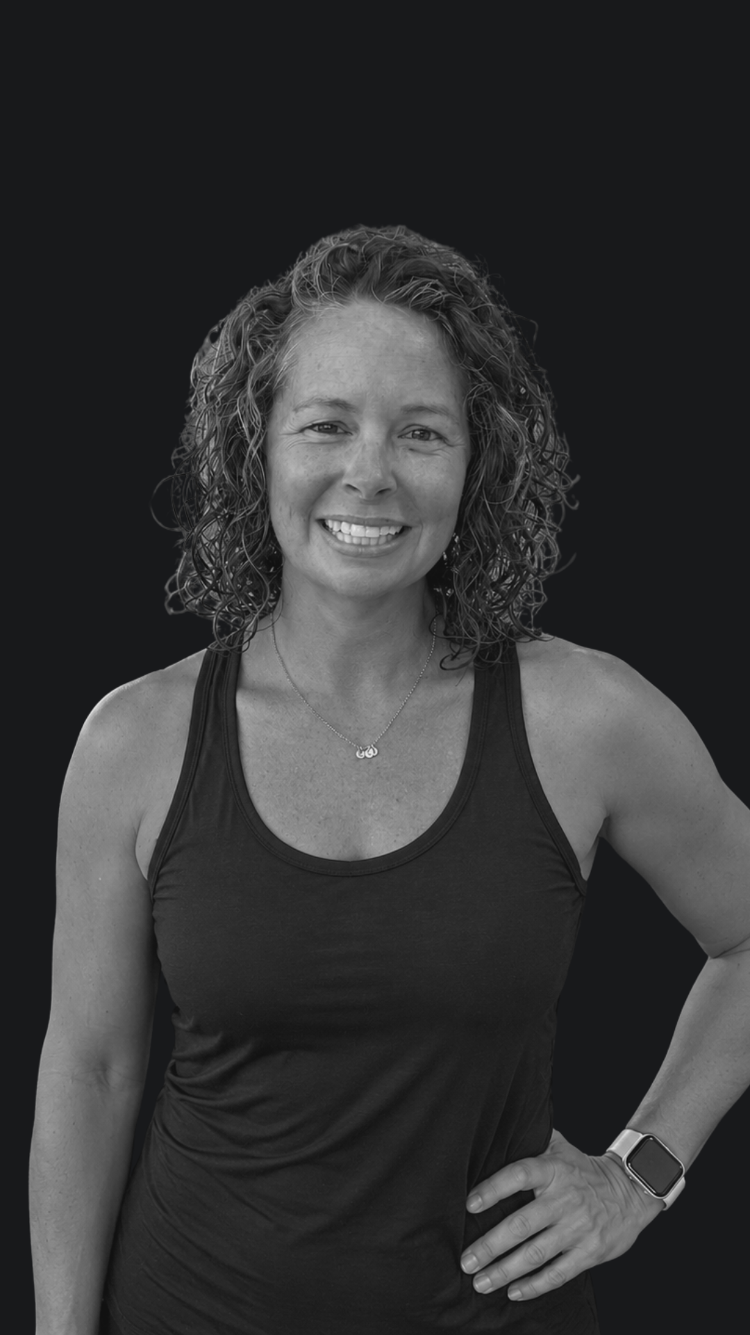 A woman with curly hair smiling, wearing a tank top, a necklace, and a smartwatch, standing against a dark background.