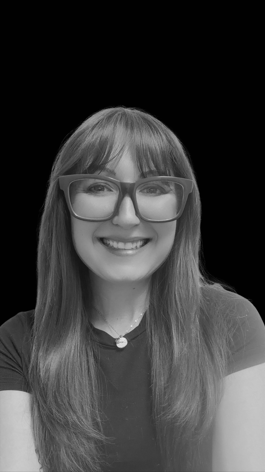 Black and white photo of a woman with long hair, wearing glasses and a necklace, smiling at the camera.