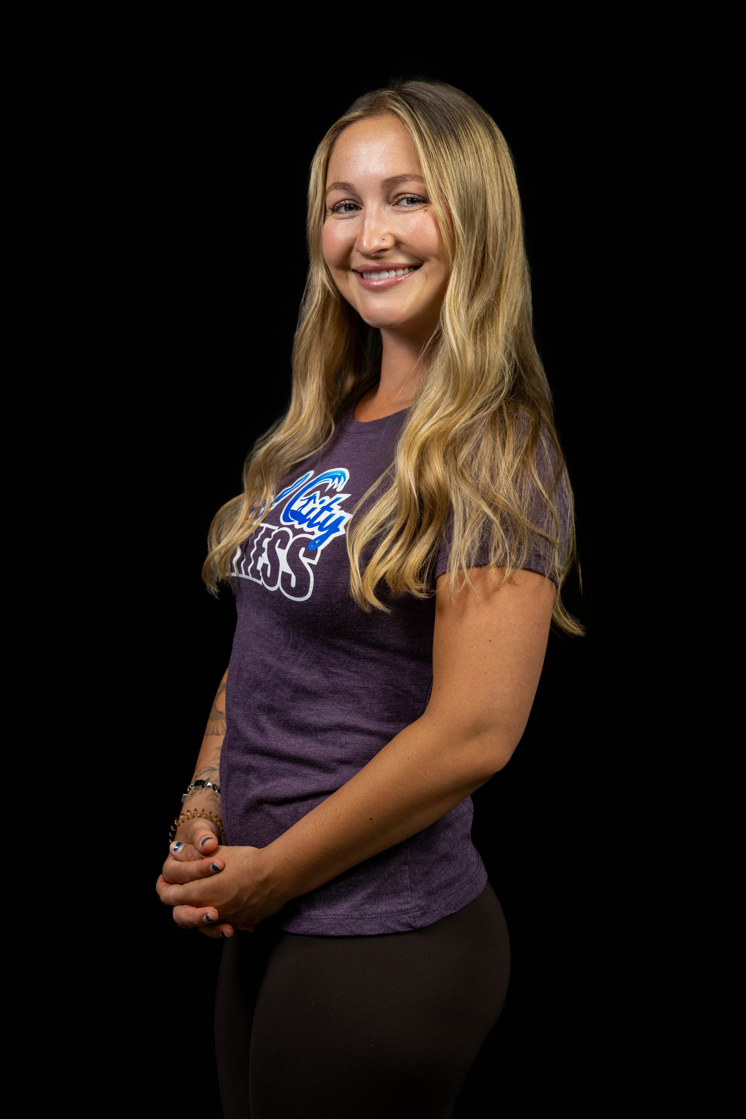 Portrait of a smiling woman with long blonde hair wearing a purple t-shirt with a logo, standing against a black background.