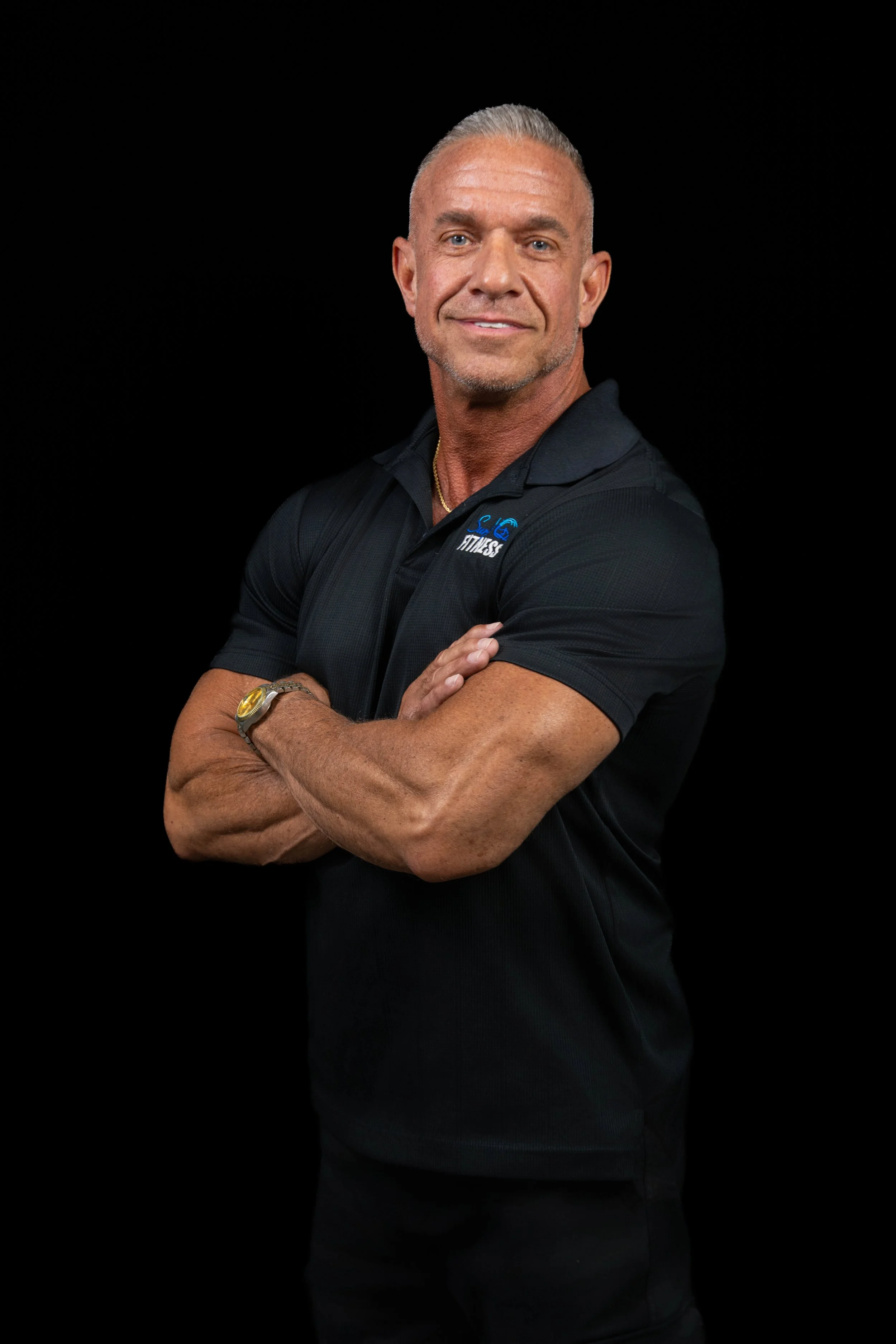 A middle-aged man with short gray hair and light skin, smiling with crossed arms, wearing a black polo shirt with a fitness logo, standing against a black background.