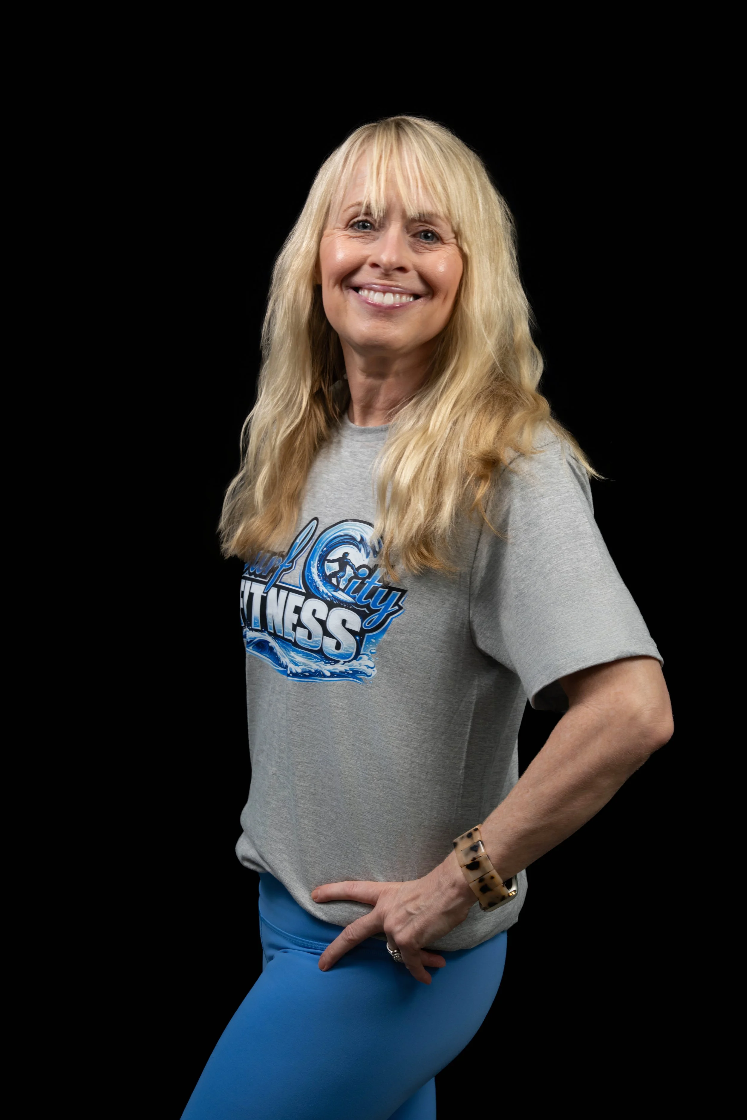 A smiling woman with long blonde hair, wearing a gray T-shirt with a fitness logo, blue leggings, and a leopard print bracelet, standing with one hand on her hip against a black background.