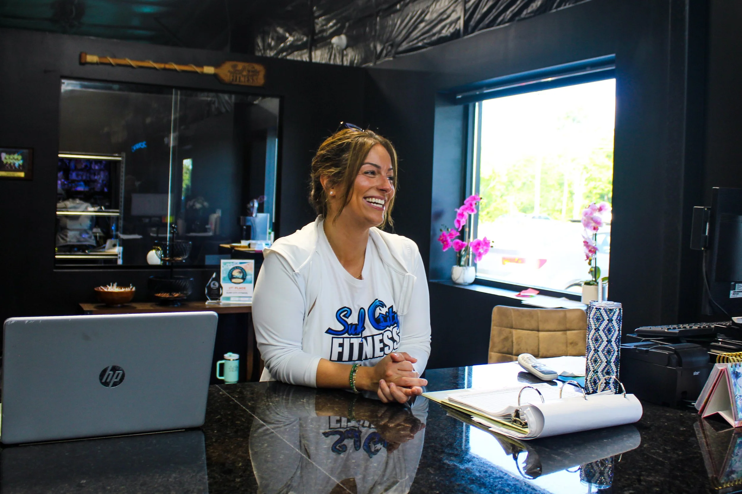 Smiling woman sitting at a reception desk with a computer, notebooks, and a vase of pink flowers in a modern, dark-colored office or reception area.