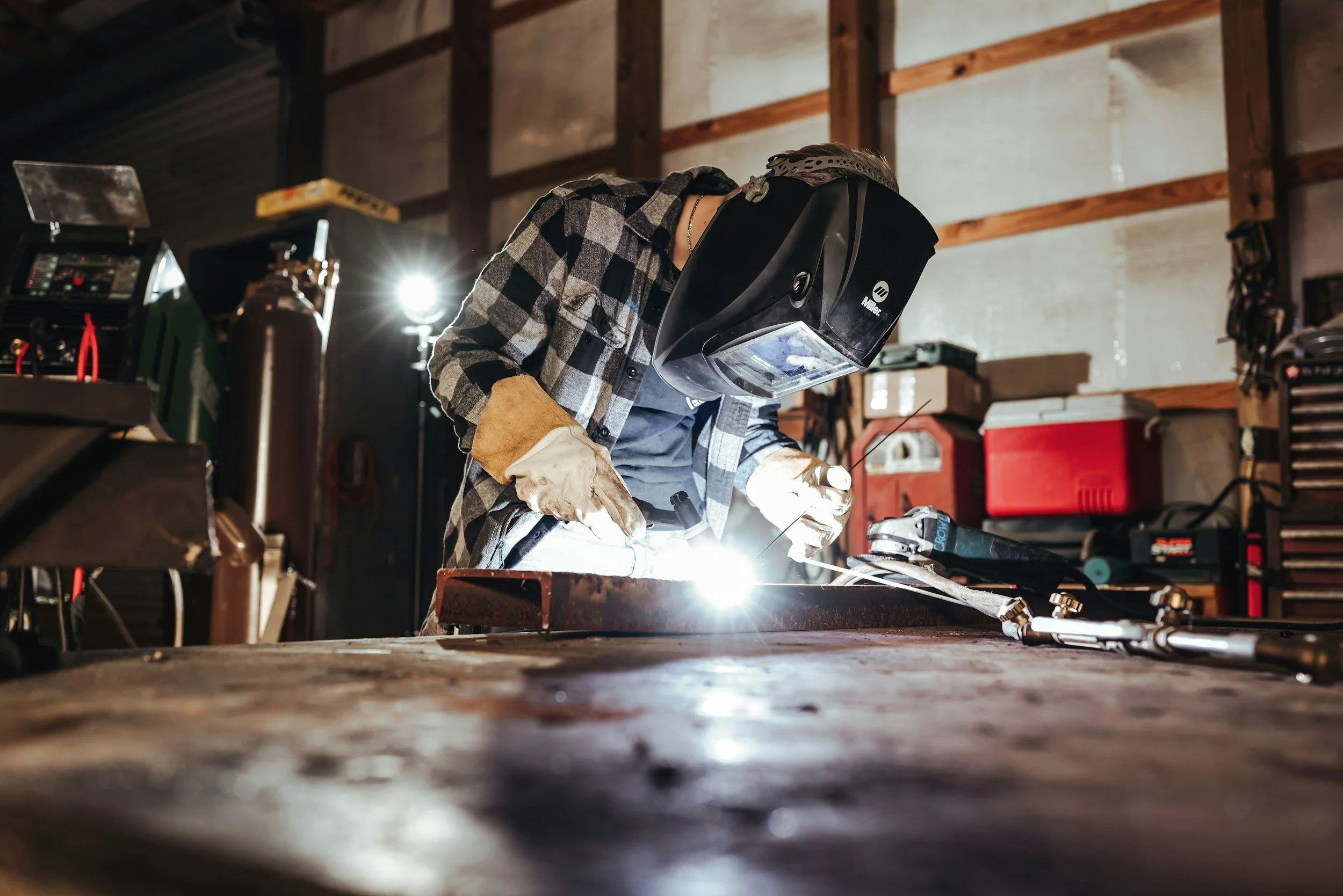 Person welding metal in a workshop, wearing protective gear including a helmet and gloves, with bright sparks flying from the welding point.