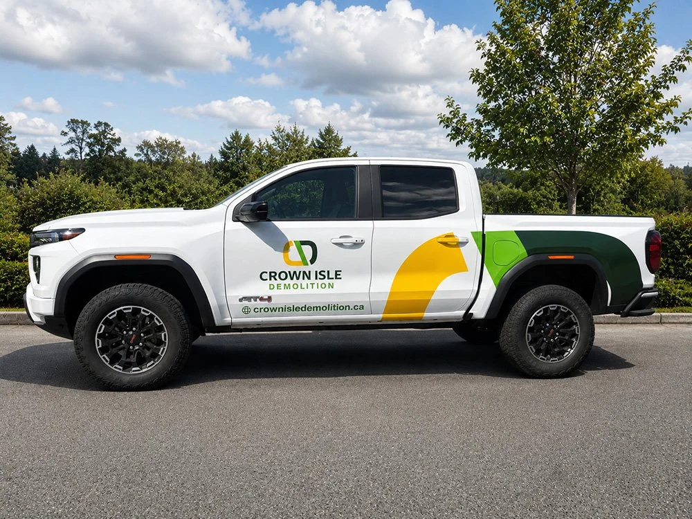 White pickup truck with Crown Isle Demolition logo, a stylized yellow and green eagle head, and website URL on the side, parked on a street with trees and cloudy sky in the background.