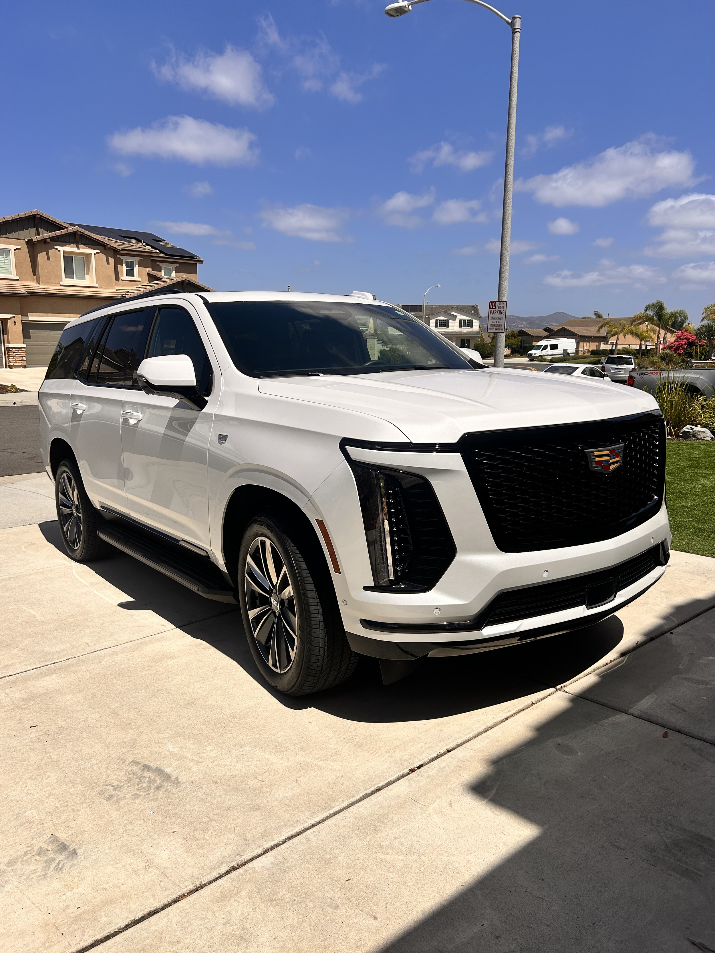 A white Cadillac SUV parked on a concrete driveway in front of a suburban house with a brown roof and some solar panels, under a blue sky with scattered clouds.