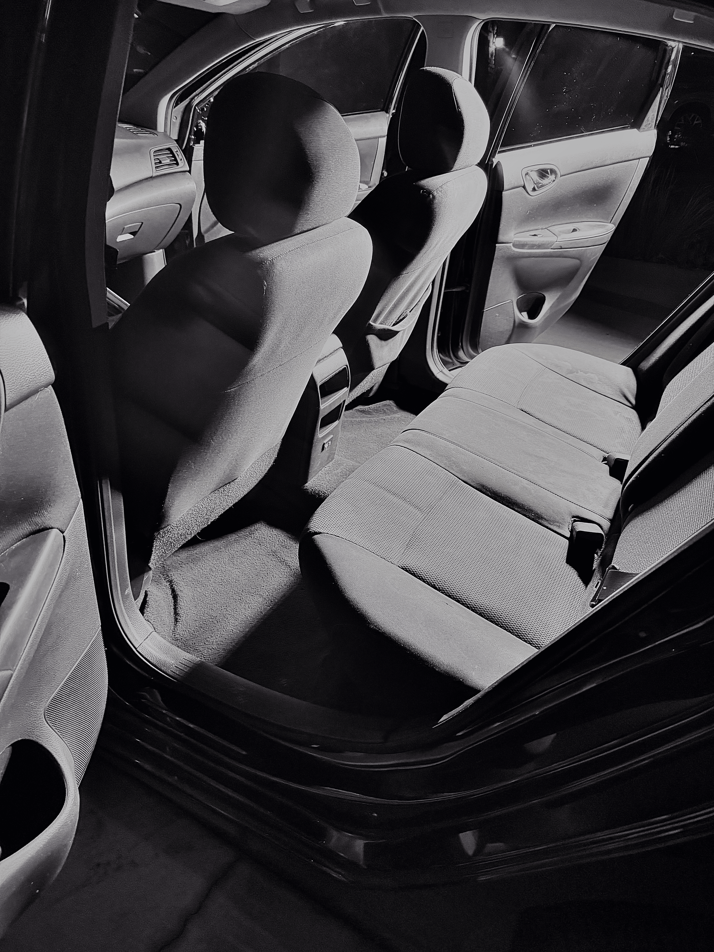 Black and white photo of the interior of a car, focusing on the backseat with fabric upholstery, front seats, and door panels visible.