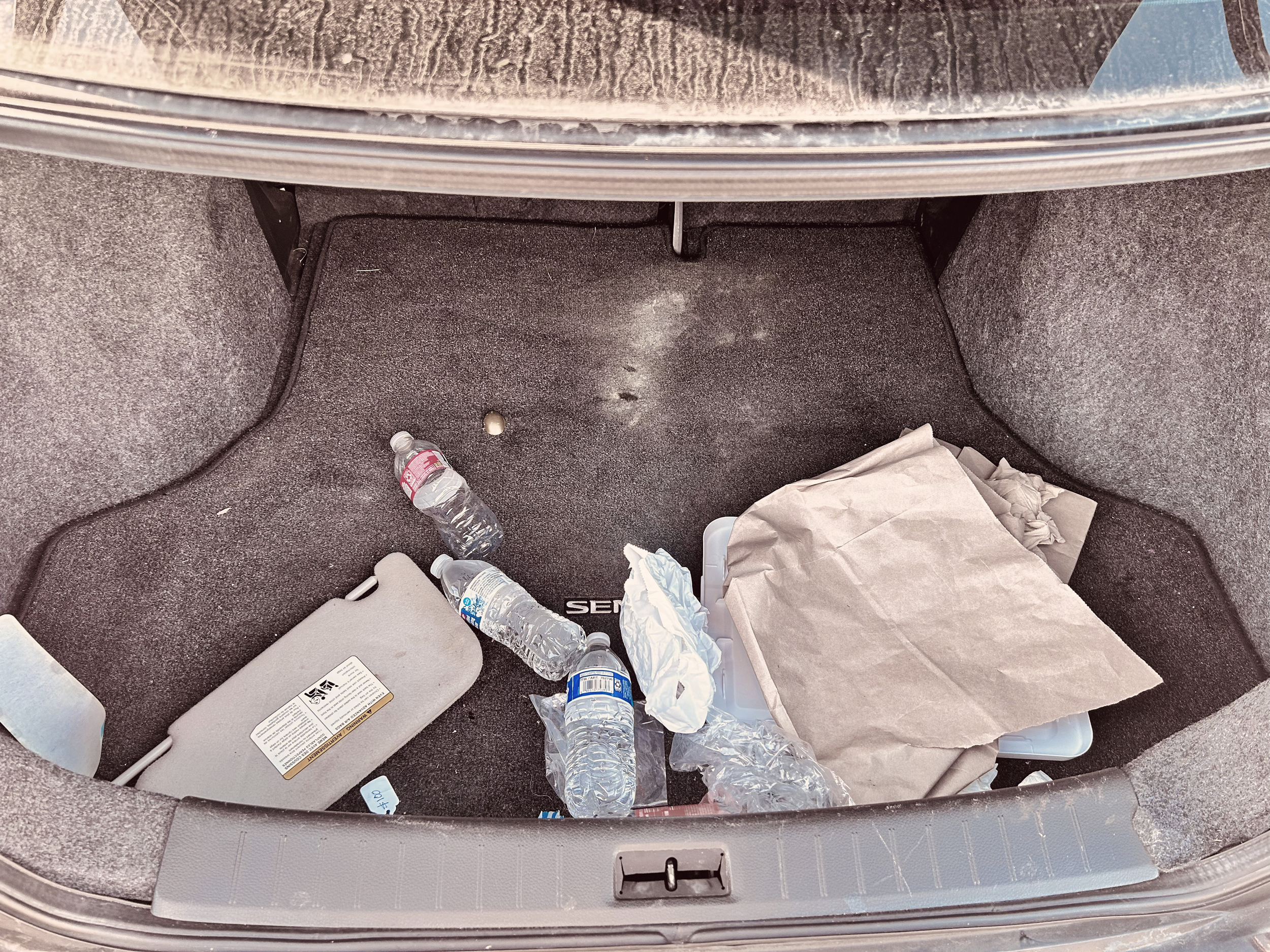 The trunk of a car with two water bottles, paper, and trash scattered on the carpeted surface.