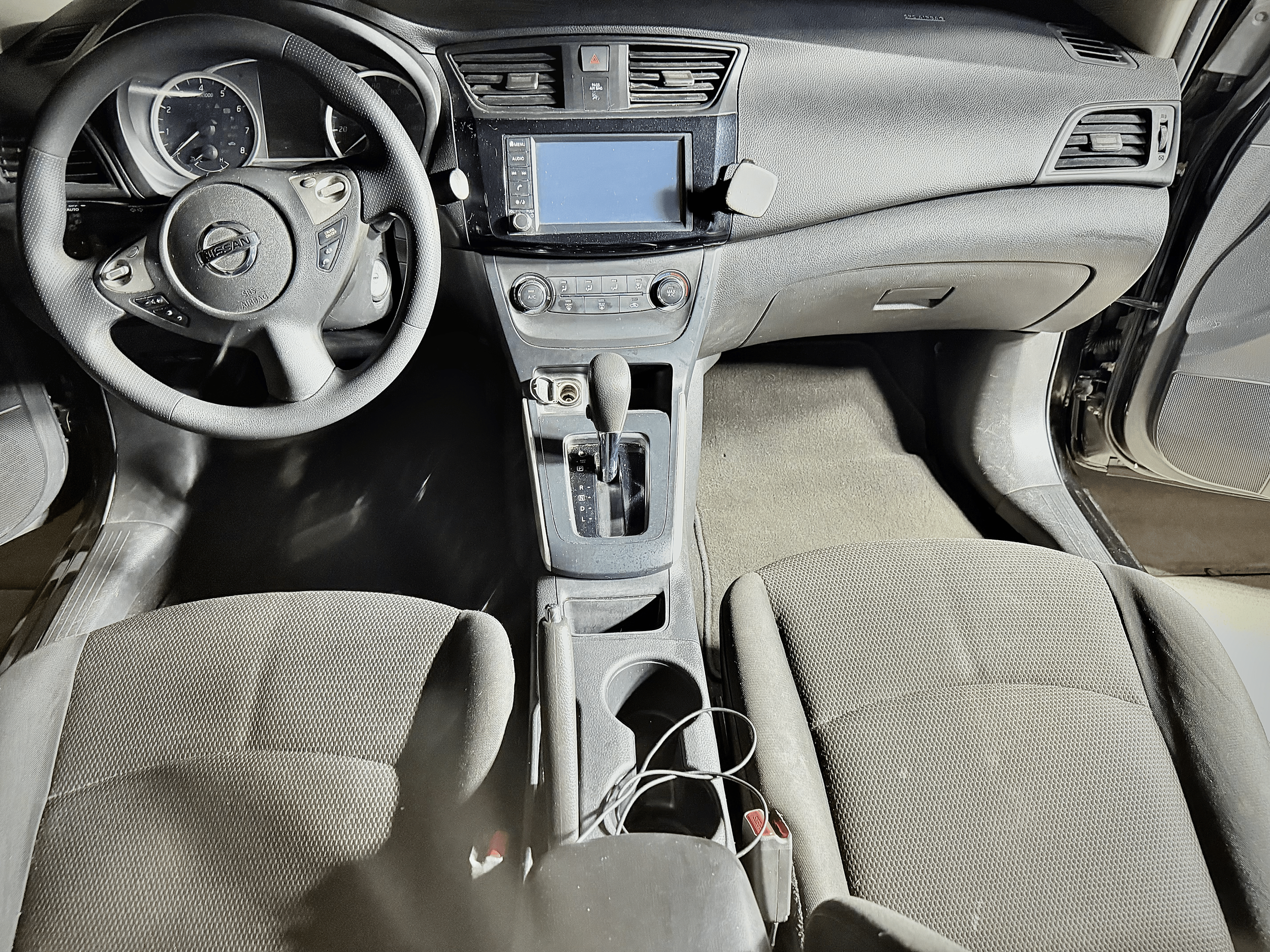 The interior of a car showing the driver's seat, steering wheel, dashboard, and center console with an infotainment screen. The dashboard includes gauges and air vents, and the gear shift is positioned in the center console. The seats are upholstered in beige fabric.