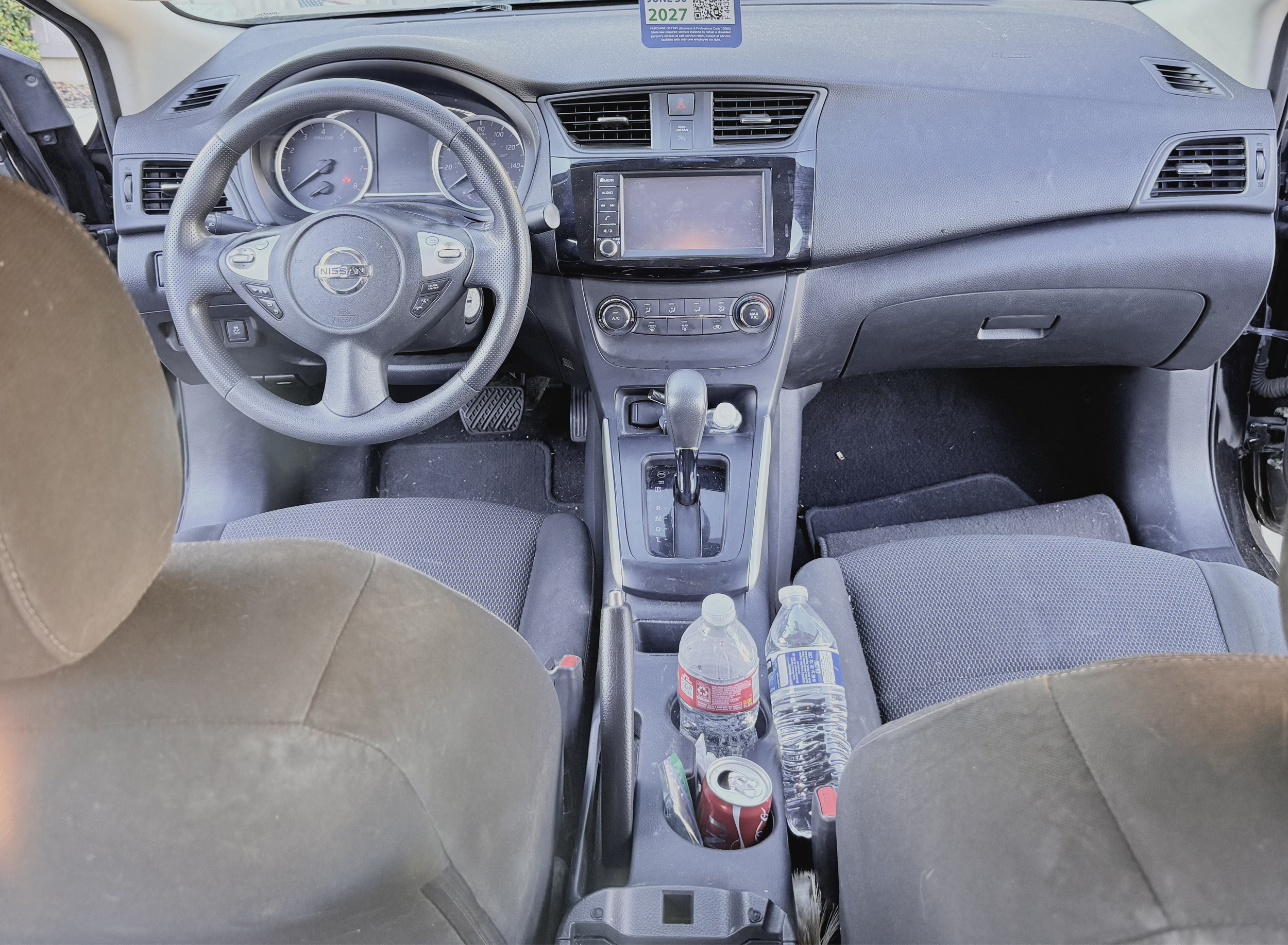 The interior of a car dashboard with a steering wheel labeled Nissan, a touchscreen display, air vents, and various control buttons. The center console has a gear shift, some bottles, and cans.