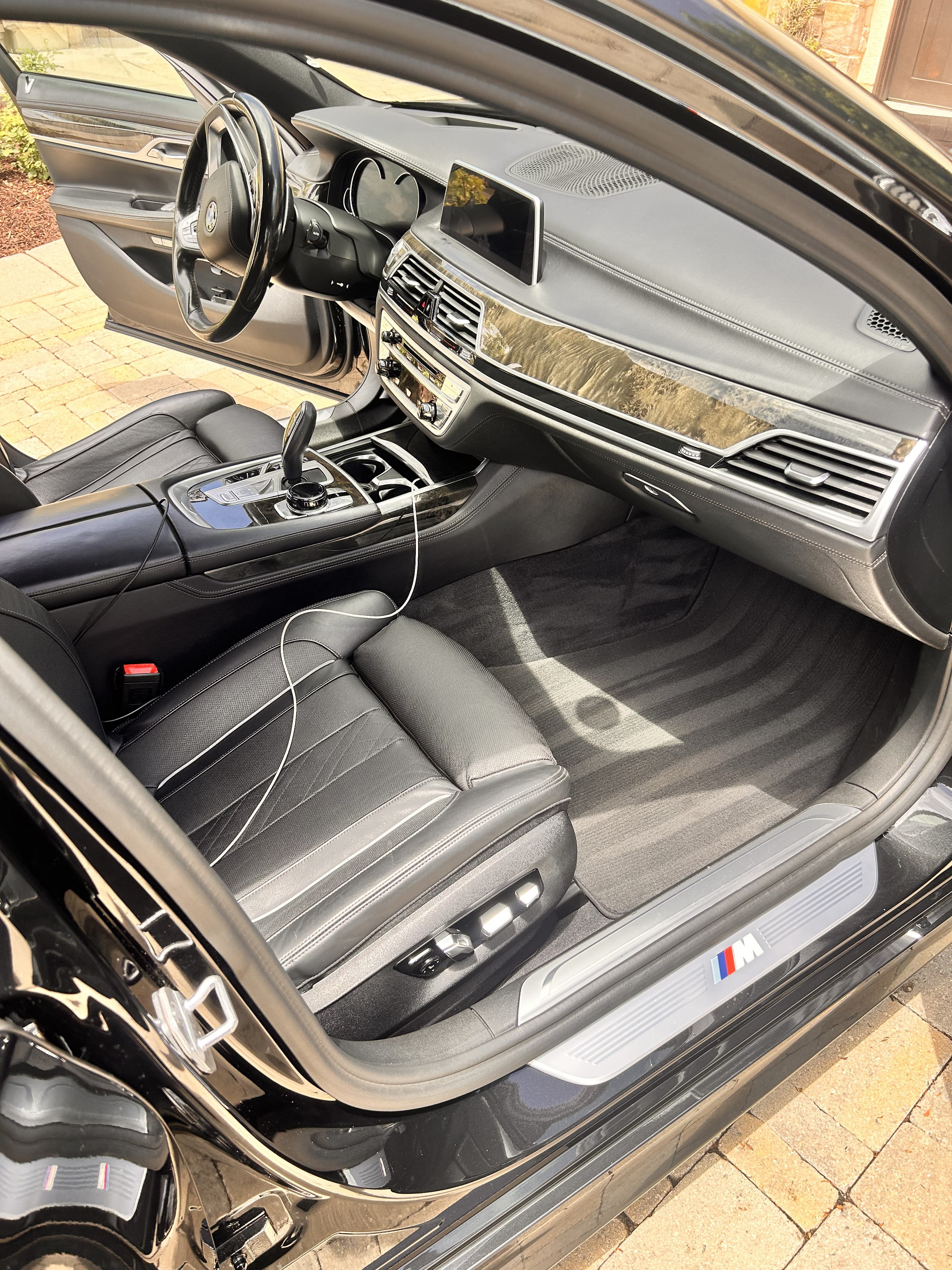 Interior of a black BMW M car, showing the driver's seat, steering wheel, dashboard, center console, and open door with a driveway and stone pathway outside.