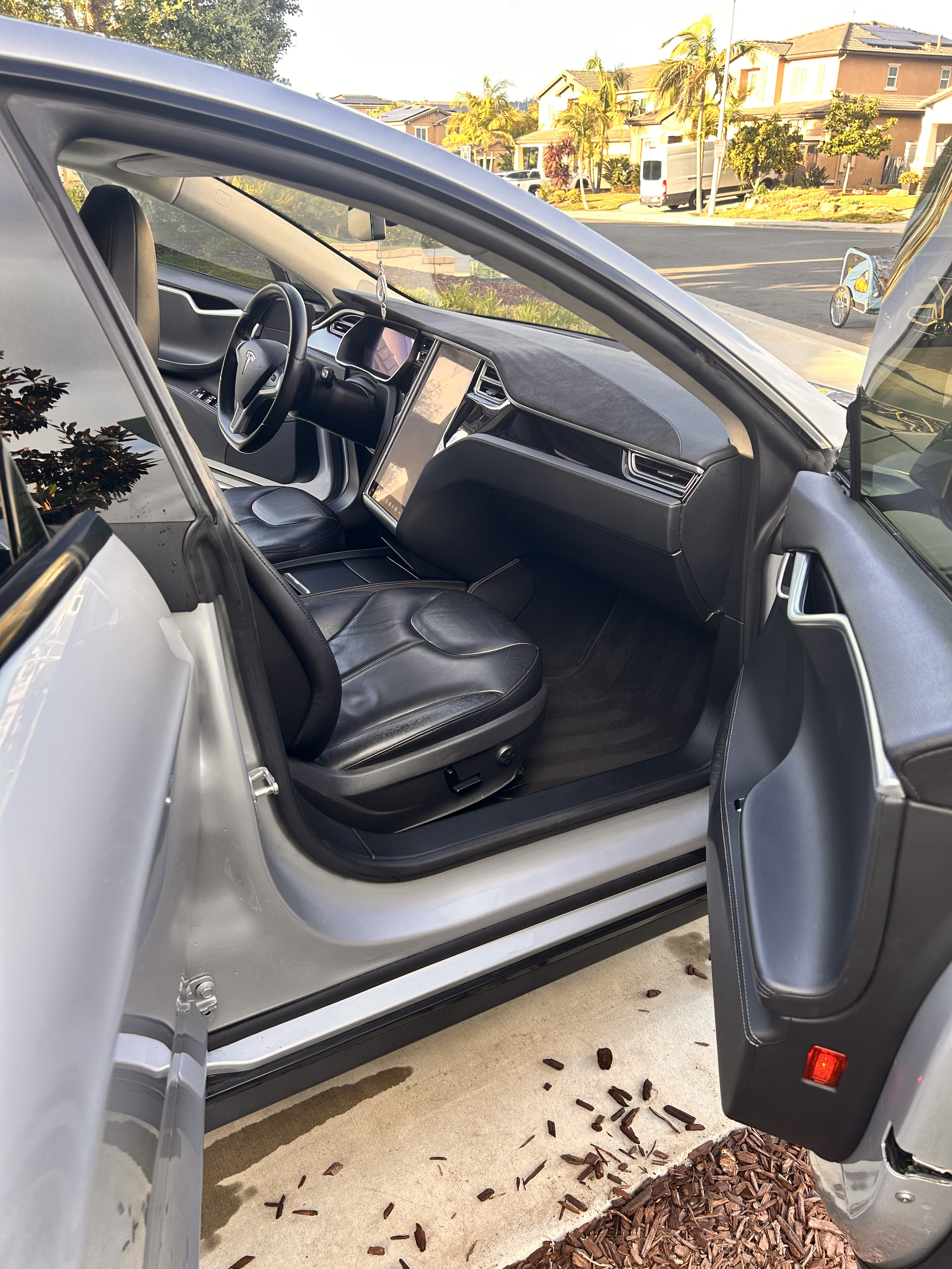 Inside of a Tesla electric vehicle parked on a driveway, with the door open, showing the black leather seats and digital dashboard screen, with residential houses and trees in the background.