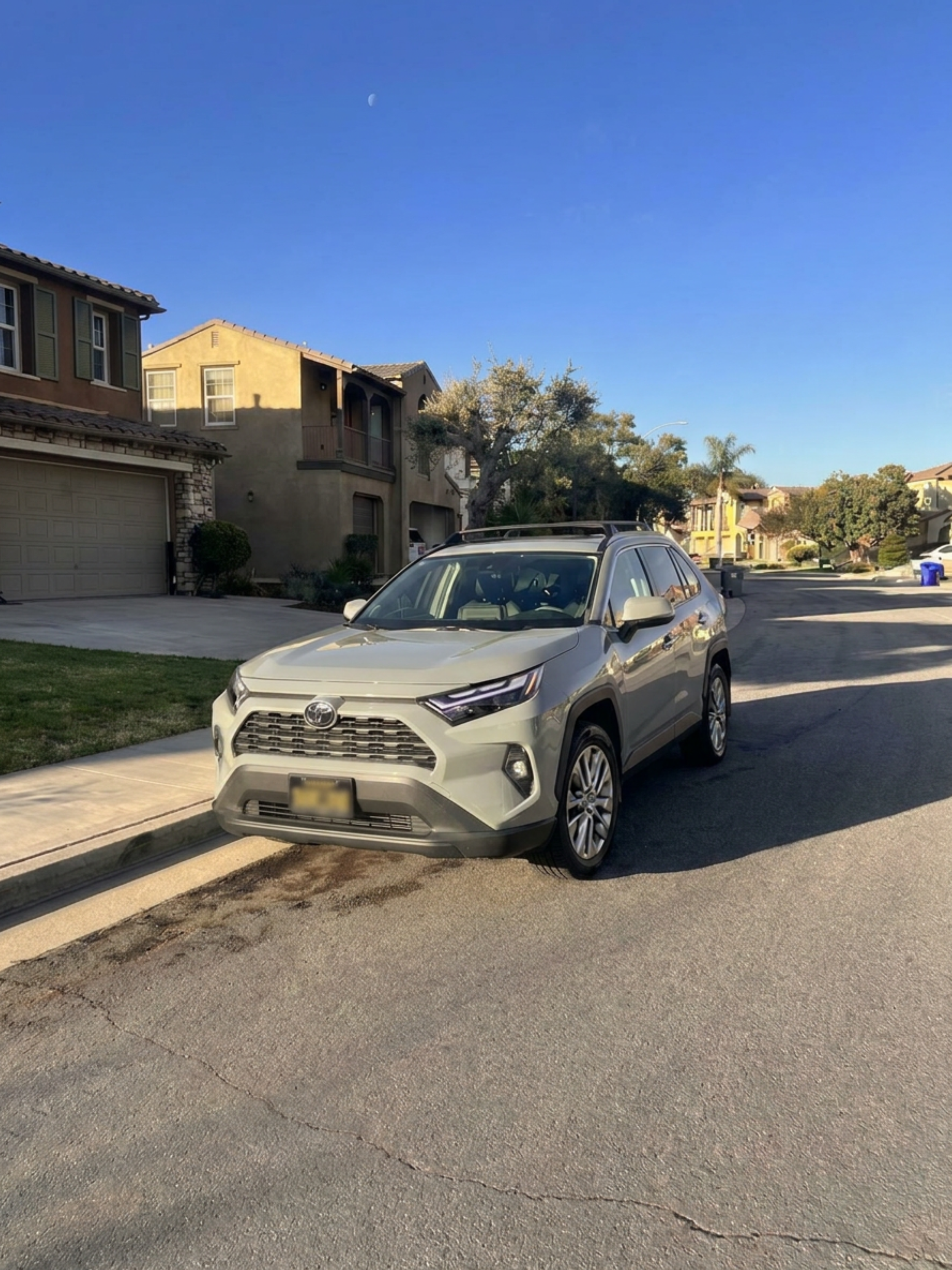 A white Toyota SUV parked on the side of a residential street in front of houses with a clear blue sky and the moon visible above.