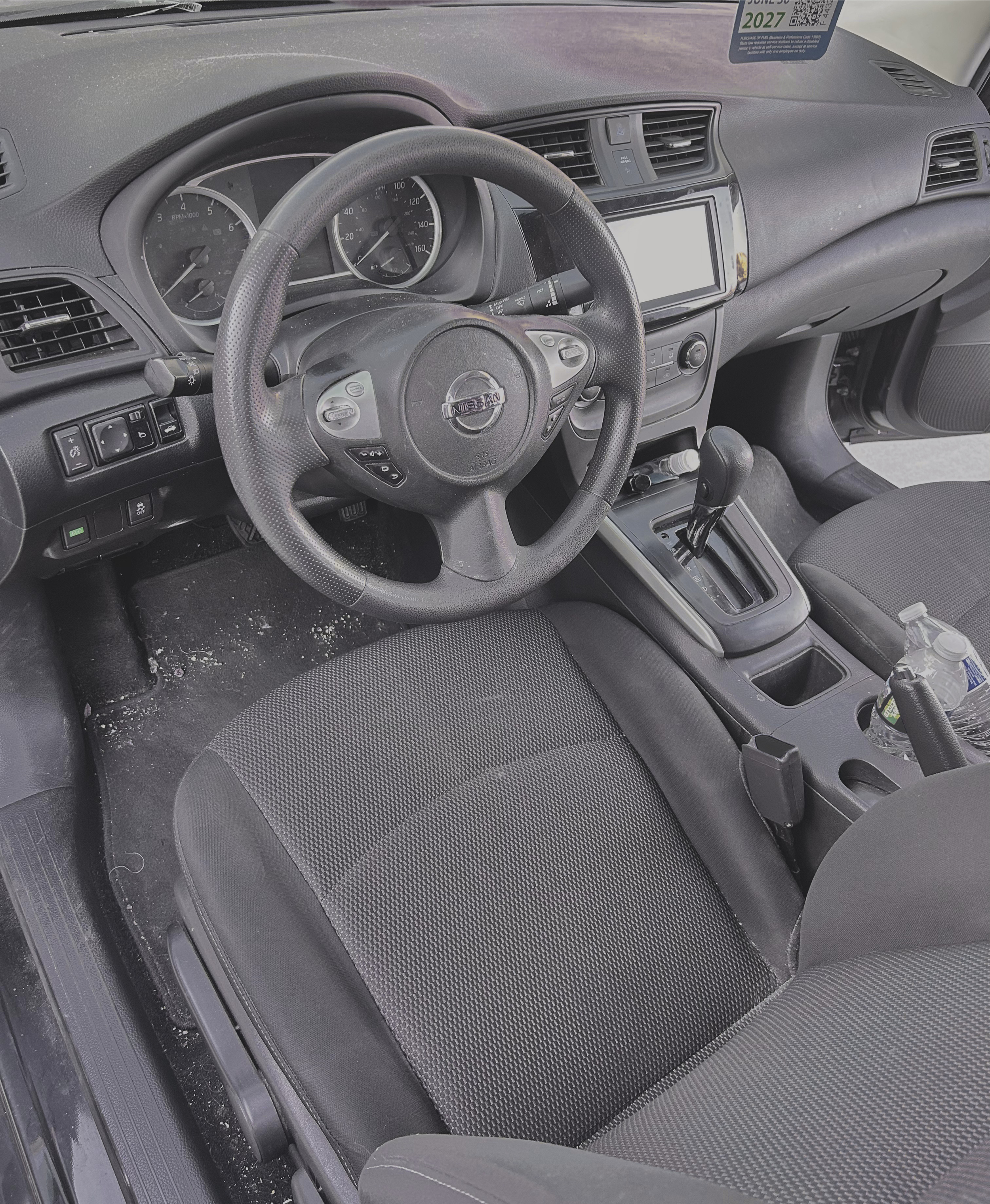 The interior of a Nissan vehicle, showing the steering wheel, dashboard with gauges, center console with a touchscreen, gear shift, and a water bottle in the cup holder. The floor is dusty with debris.