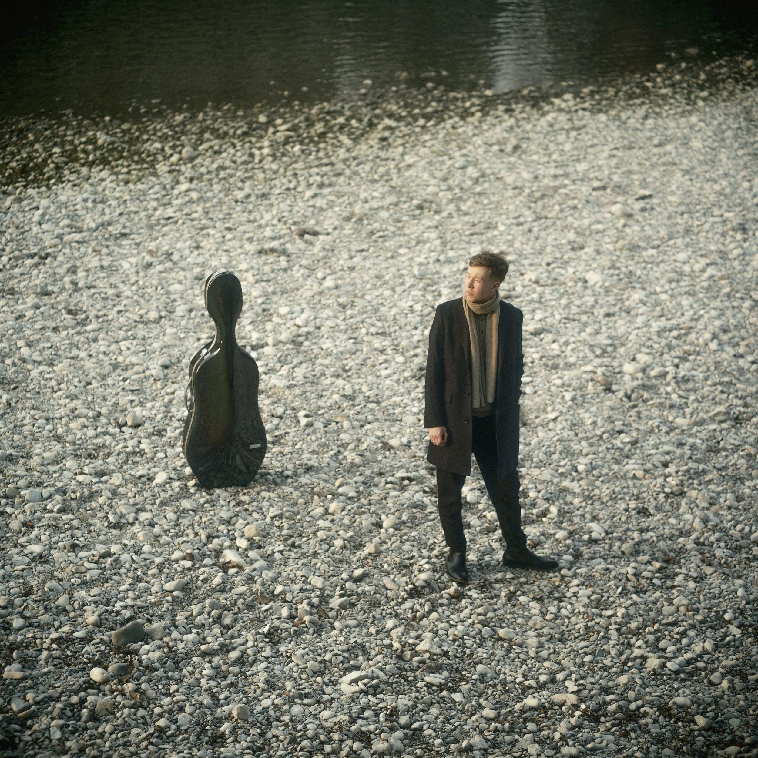 A man in a dark coat and scarf standing on a rocky beach with a guitar case nearby, facing away from the water with his head turned to the side, on a cloudy day.