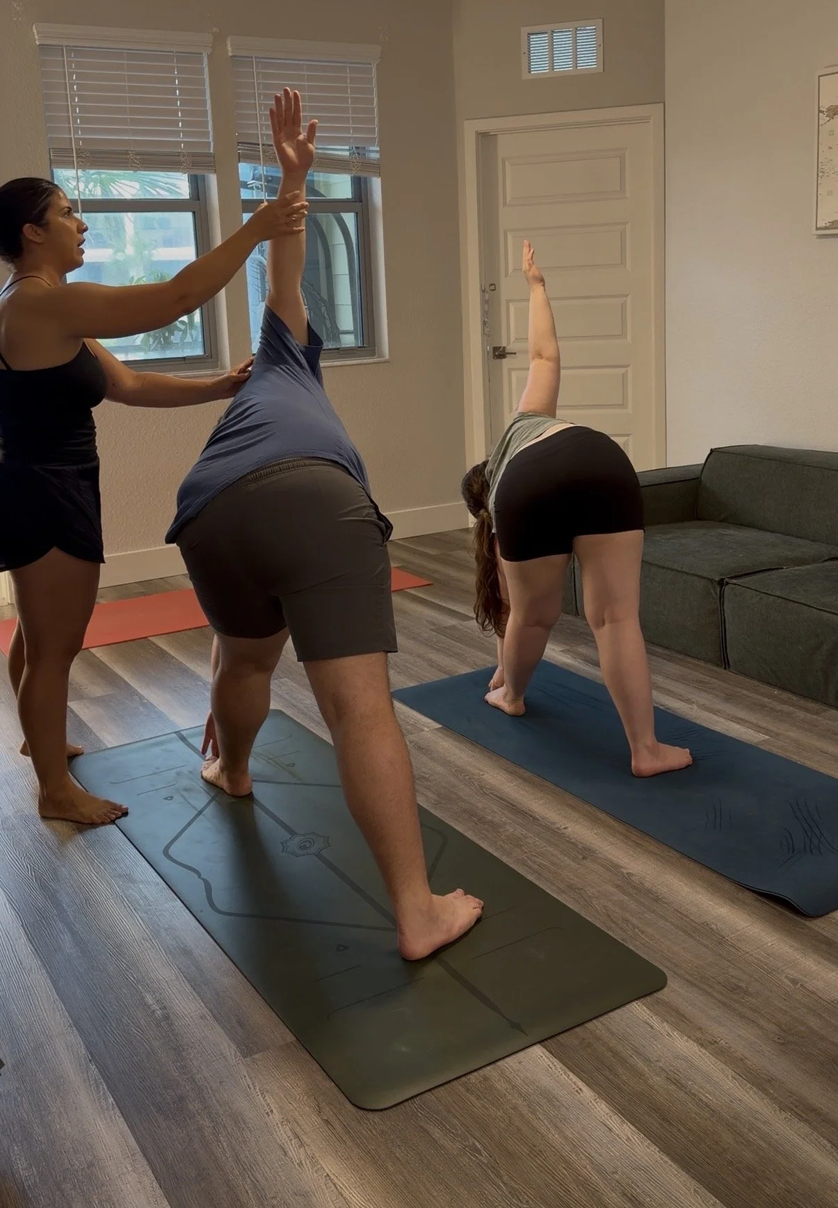Three women practicing yoga indoors on exercise mats; one woman in a downward dog pose, another supporting her from behind, and the third assisting with a stretch.