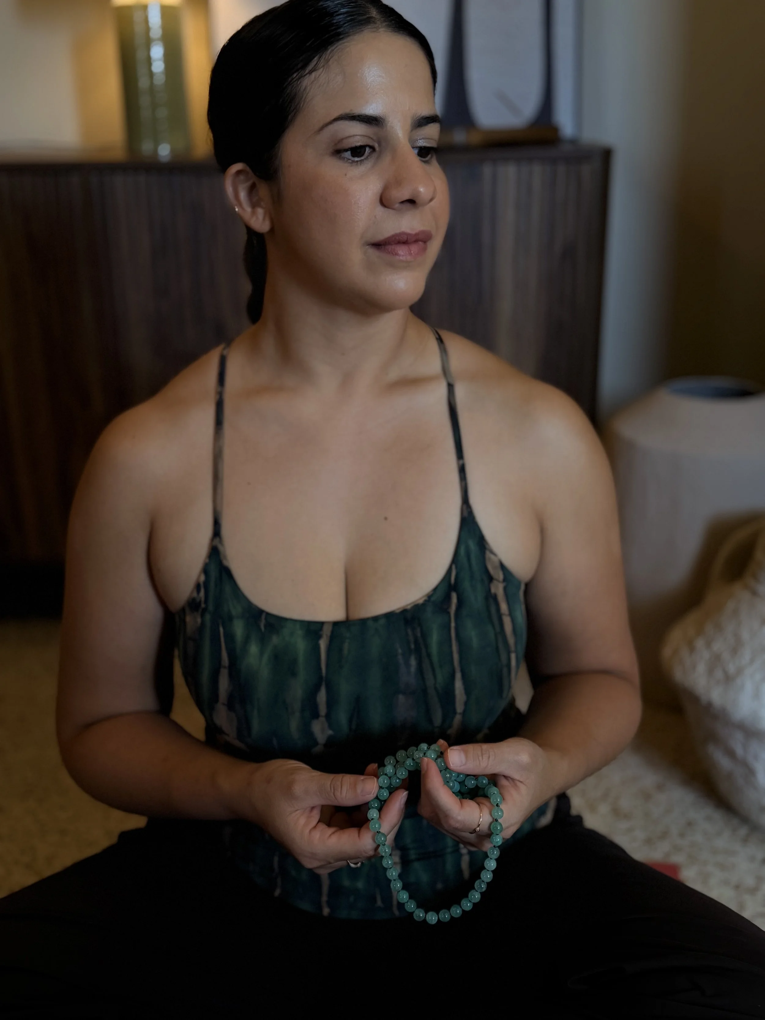 A woman with dark hair, wearing a spaghetti strap top, is sitting and holding a string of turquoise beads in her hands.