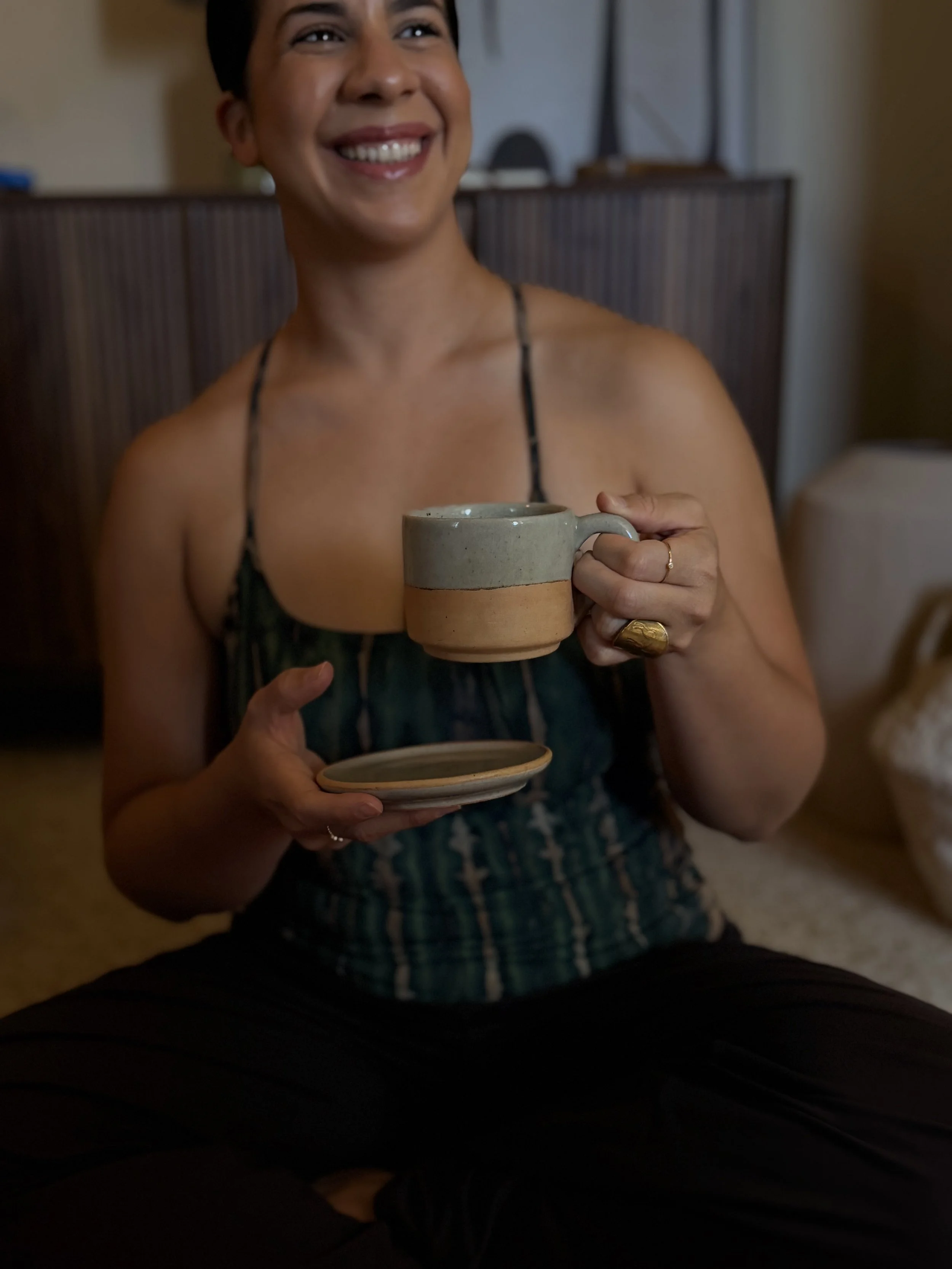 A person smiling and holding a ceramic cup and saucer, wearing a dark sleeveless top with a patterned design, sitting indoors.