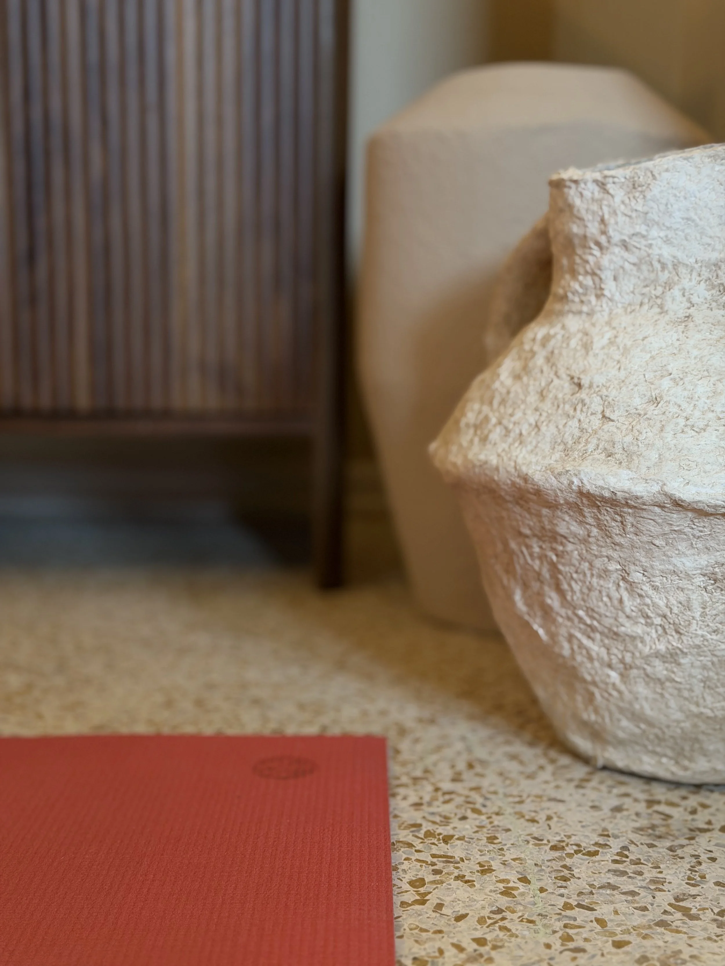 Close-up of a textured beige ceramic flowerpot on a speckled terrazzo floor with a brown wooden cabinet and a non-illuminated cream-colored lamp in the background.