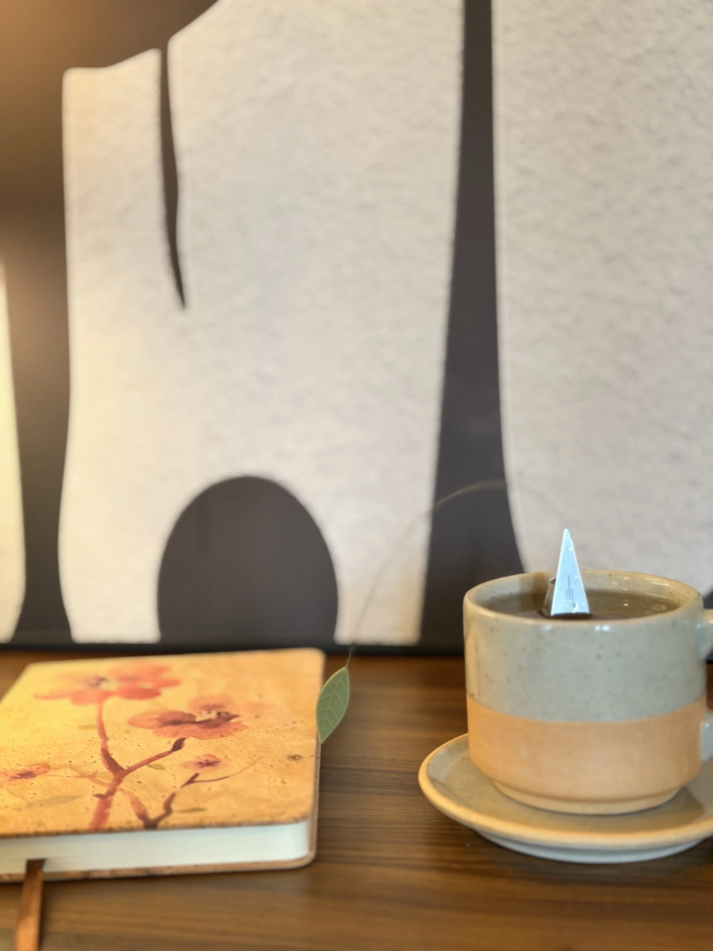 A beige coffee cup with a white triangular tea bag inside, placed on a matching beige saucer, on a wooden table with a book featuring cherry blossom artwork and Japanese writing, and a painting with black silhouettes of trees in the background.
