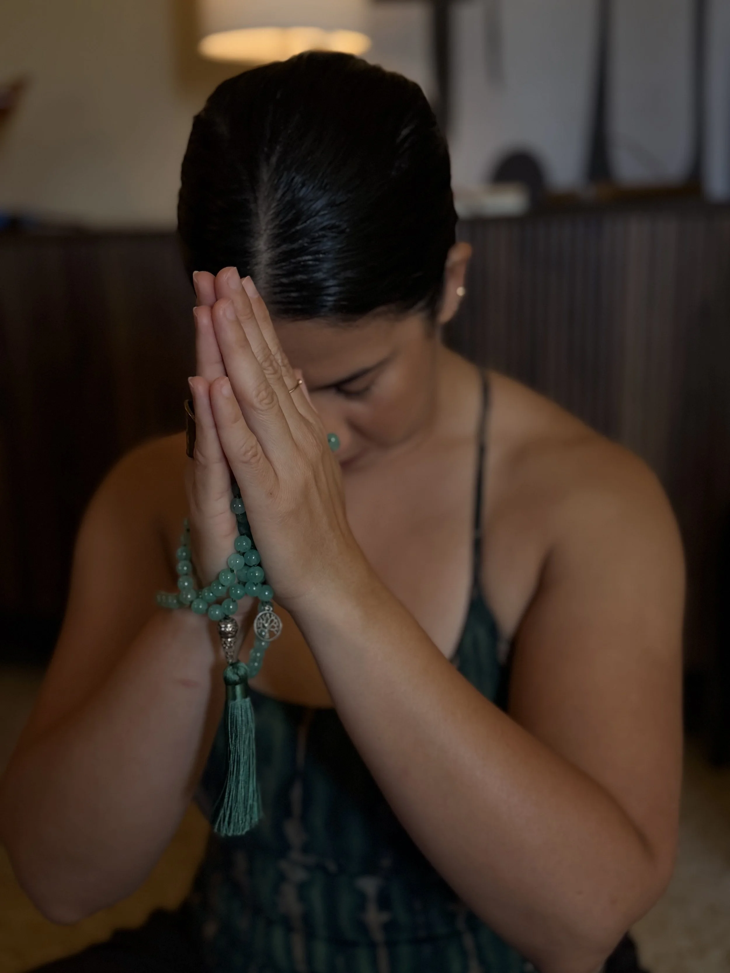 A woman with dark hair pulled back, wearing a sleeveless dark dress, has her hands pressed together in a prayer-like pose with her head bowed down, wearing rings and beaded bracelets, in a dimly lit room.