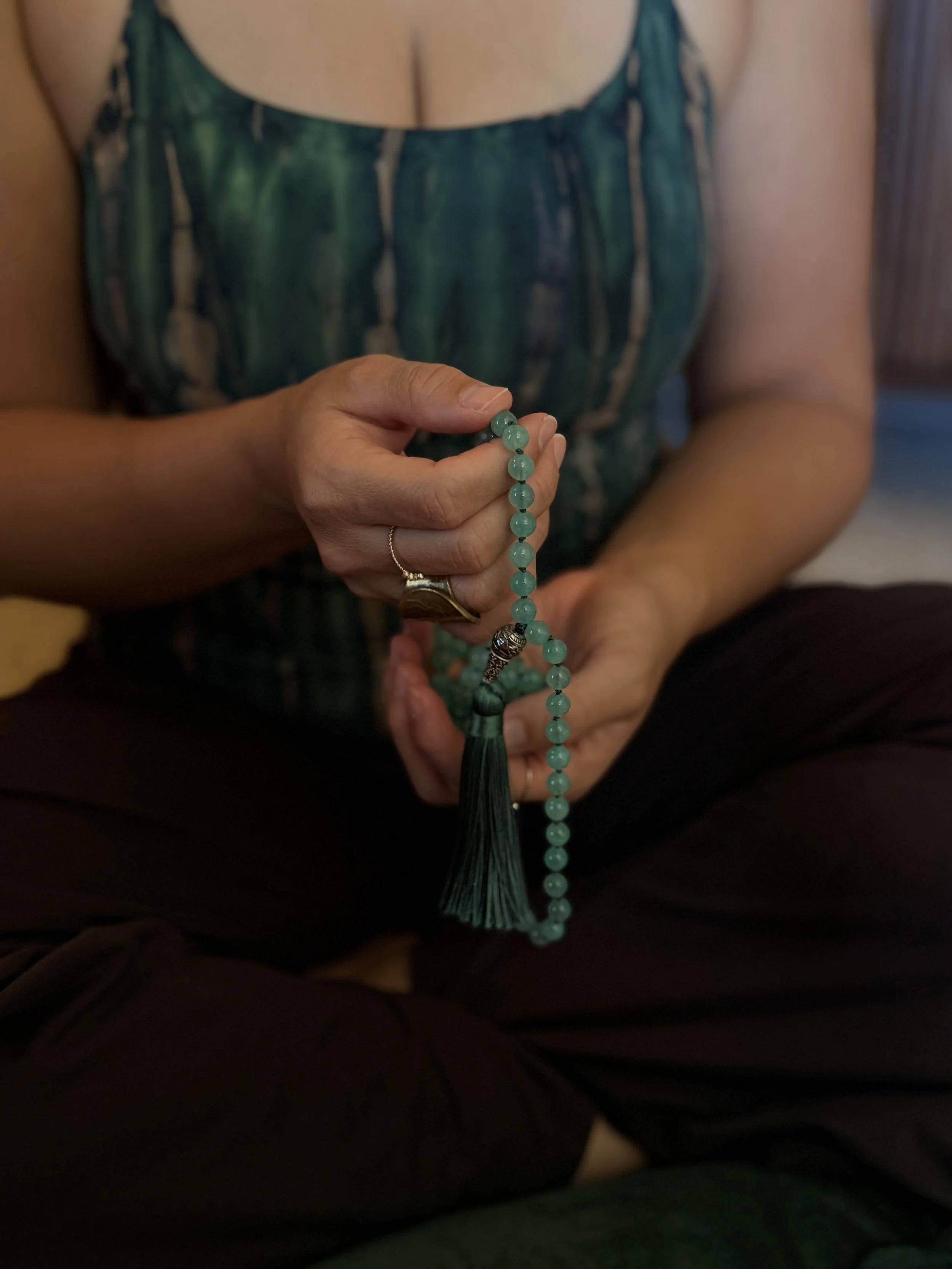 A person holding a string of turquoise prayer beads with a tassel, wearing a ring on one finger and a skirt with a green and blue pattern.