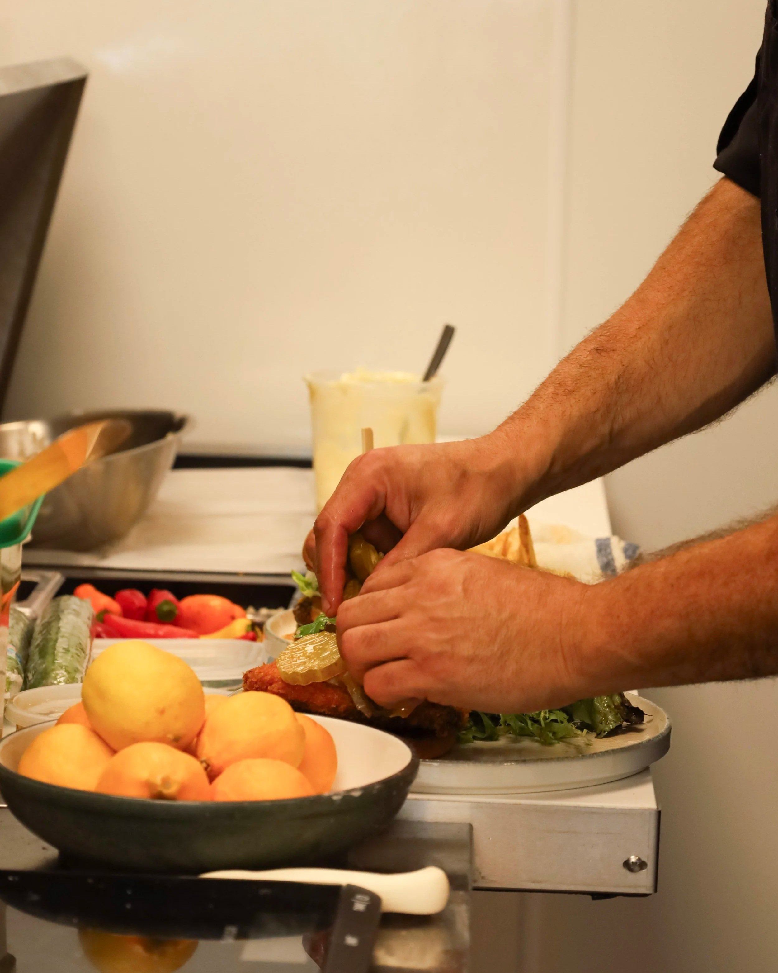 Person preparing a sandwich or wrap with various fresh ingredients on a kitchen counter, including a bowl of peaches, colorful vegetables, and a glass of dip or sauce in the background.