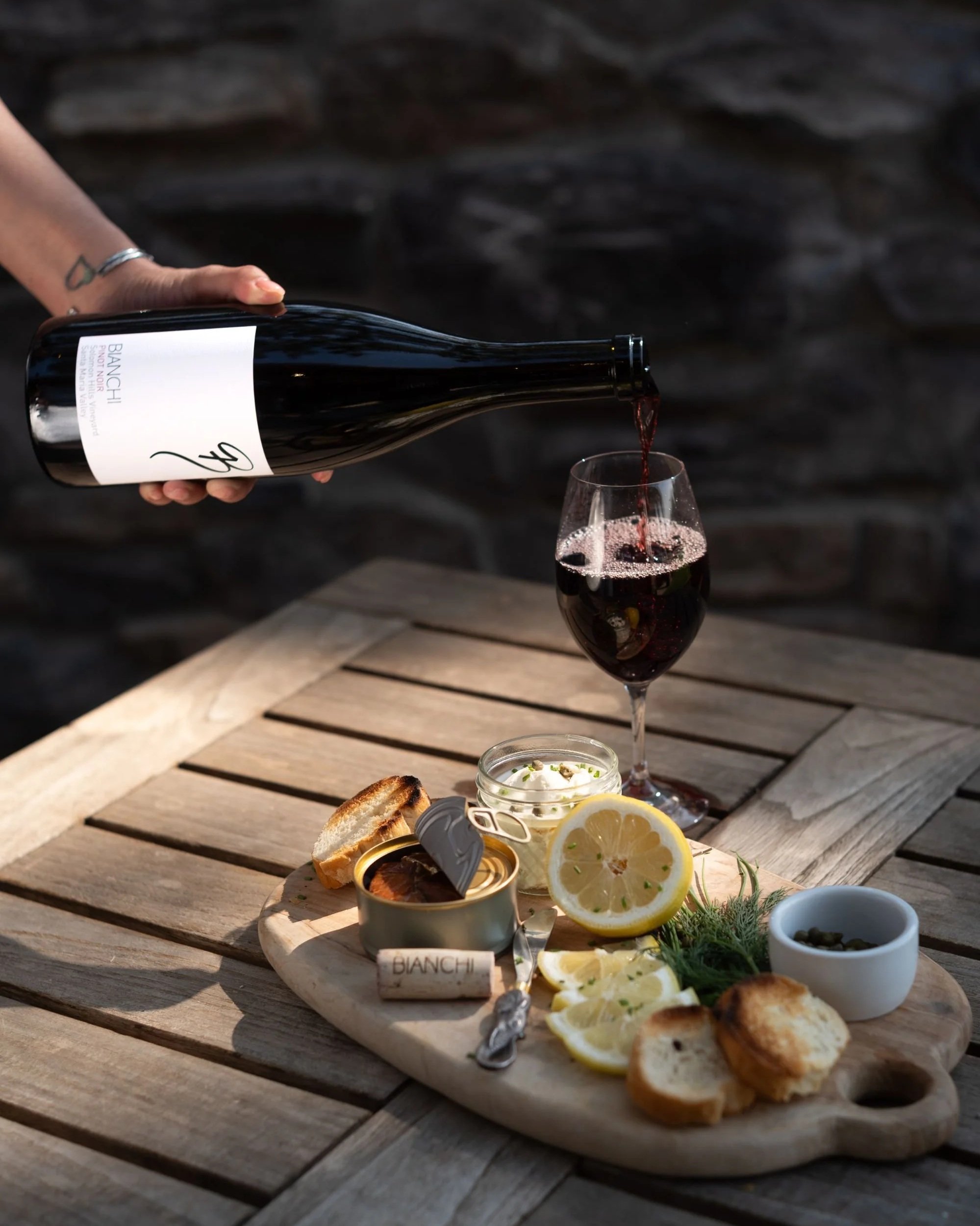 A hand pouring red wine from a bottle into a wine glass on a wooden table, with a cheese platter featuring lemon slices, bread, and condiments.