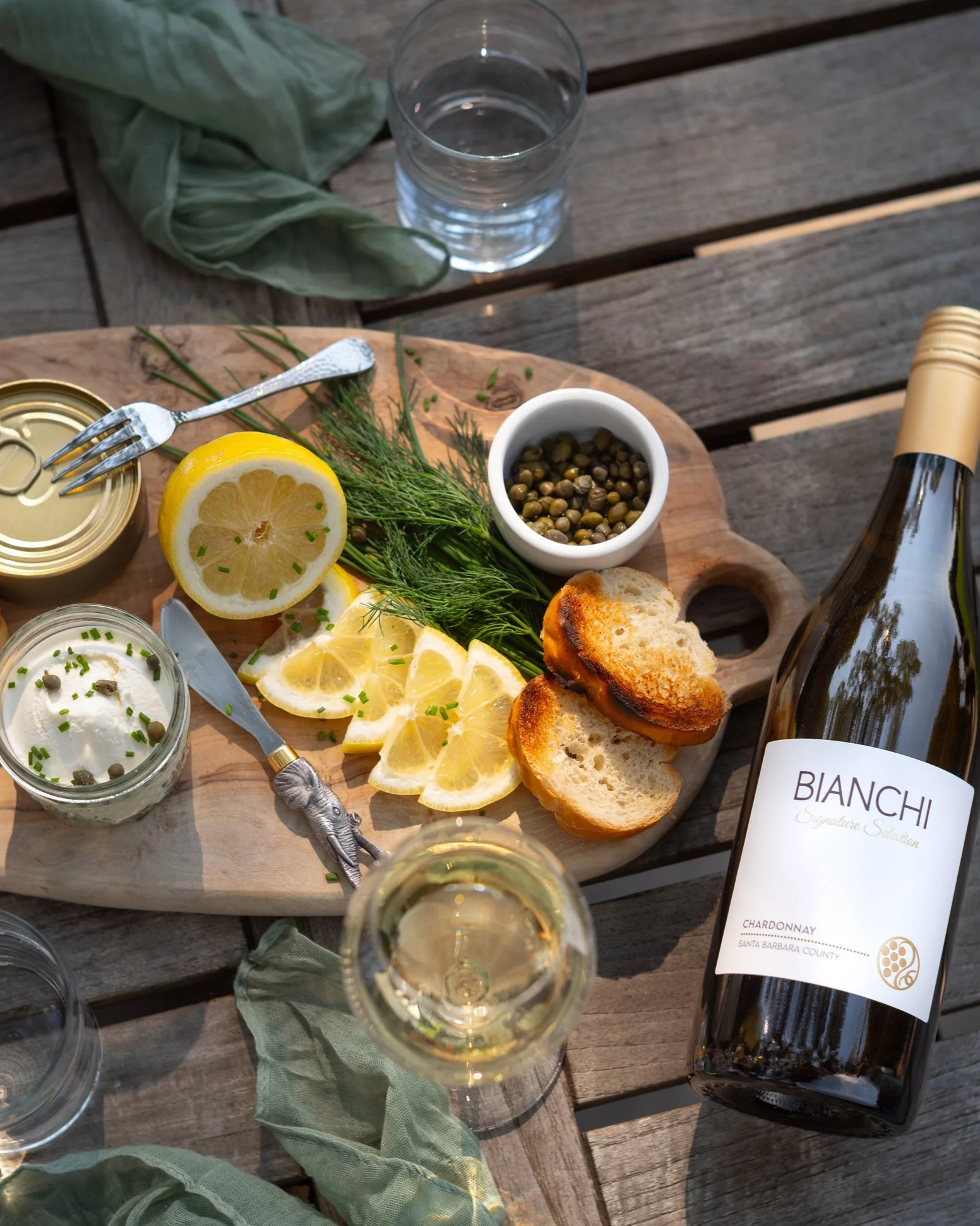 A wooden serving board with sliced lemon, fresh herbs, a small dish of capers, slices of toasted bread, and jars of spread or cheese. There is also a glass of white wine, a bottle of Chardonnay wine labeled 'Bianchi,' and a glass of water on a wooden table.