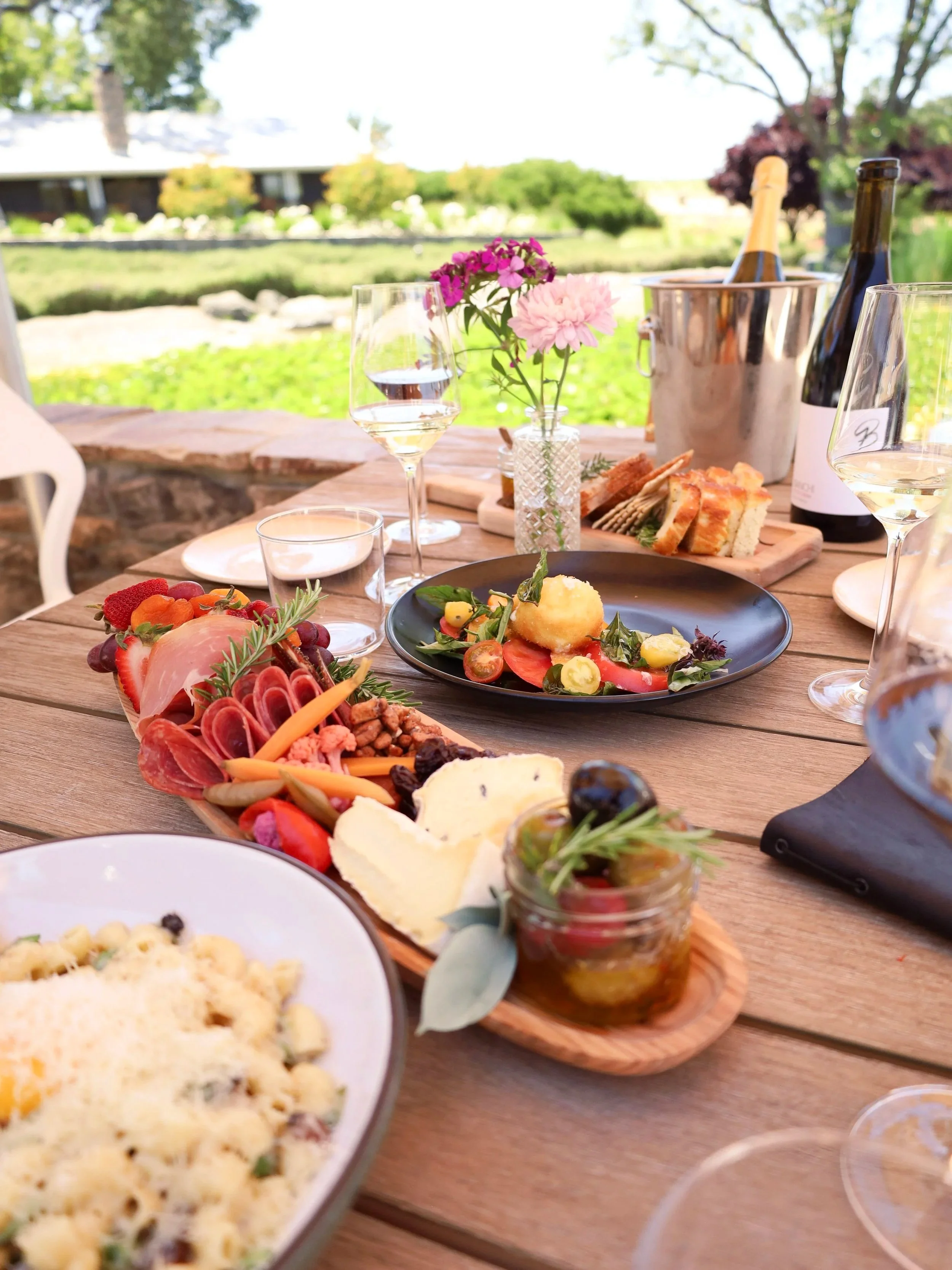 Outdoor table set with various dishes, glasses of white wine, a vase with pink flowers, bread, cheese, and charcuterie against a garden background.