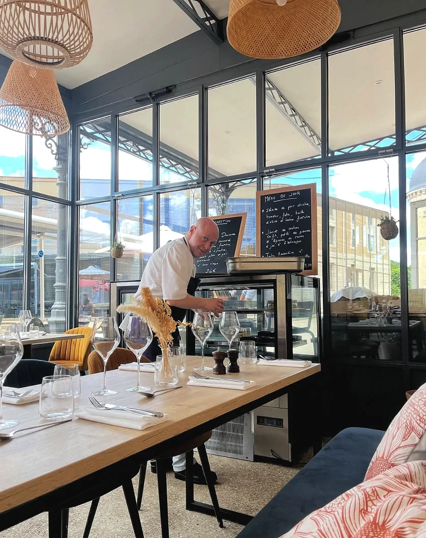 Un chef souriant prépare des plats dans un restaurant élégant avec grande baie vitrée, tables dressées avec des verres à vin, assiettes, et décorations comme des petits paniers suspendus et des fleurs séchées.