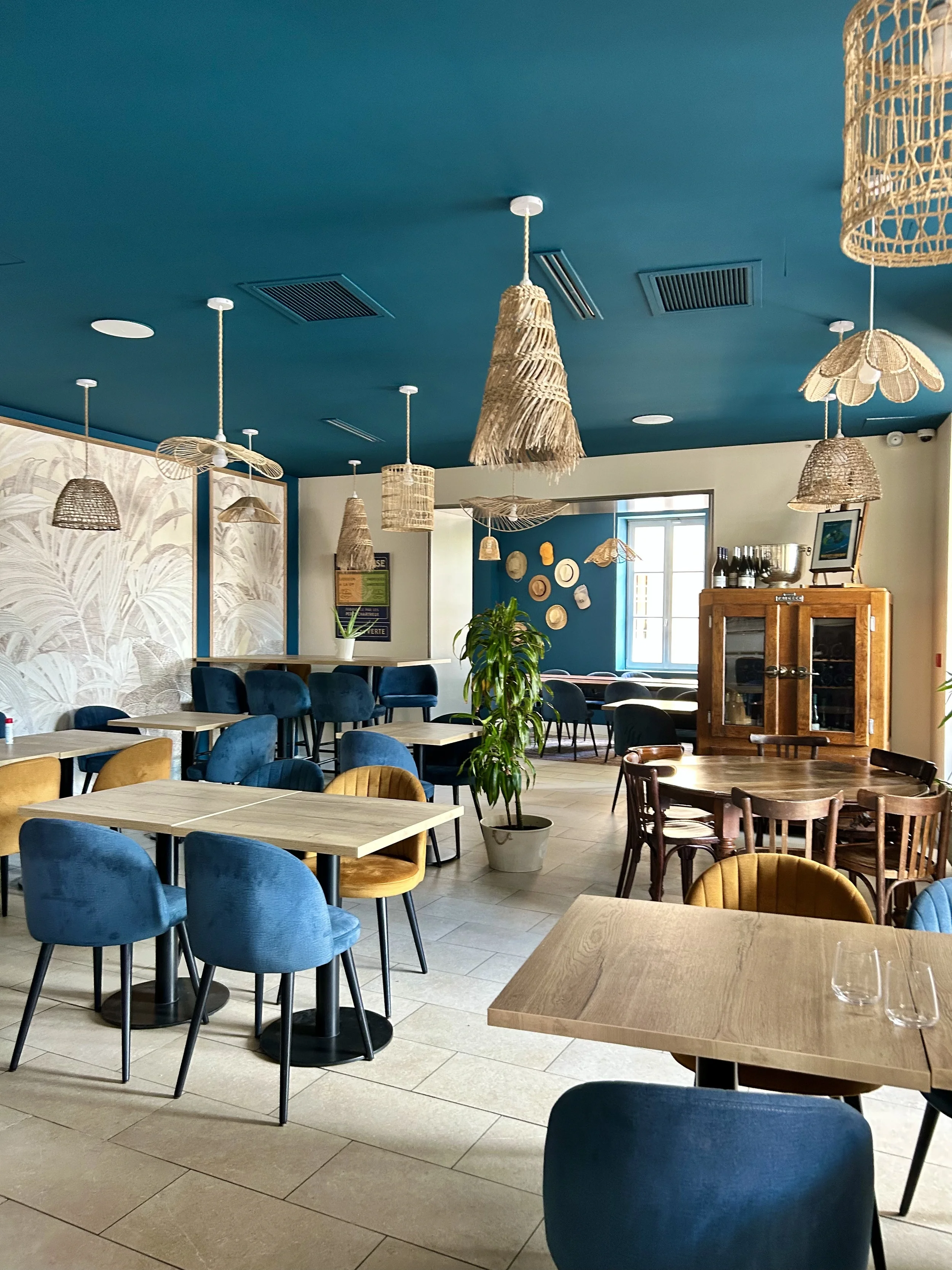 Intérieur d'un restaurant ou café avec des tables en bois, des chaises bleues et en bois, des suspensions en osier et des plantes vertes, avec une fenêtre laissant entrer la lumière naturelle.