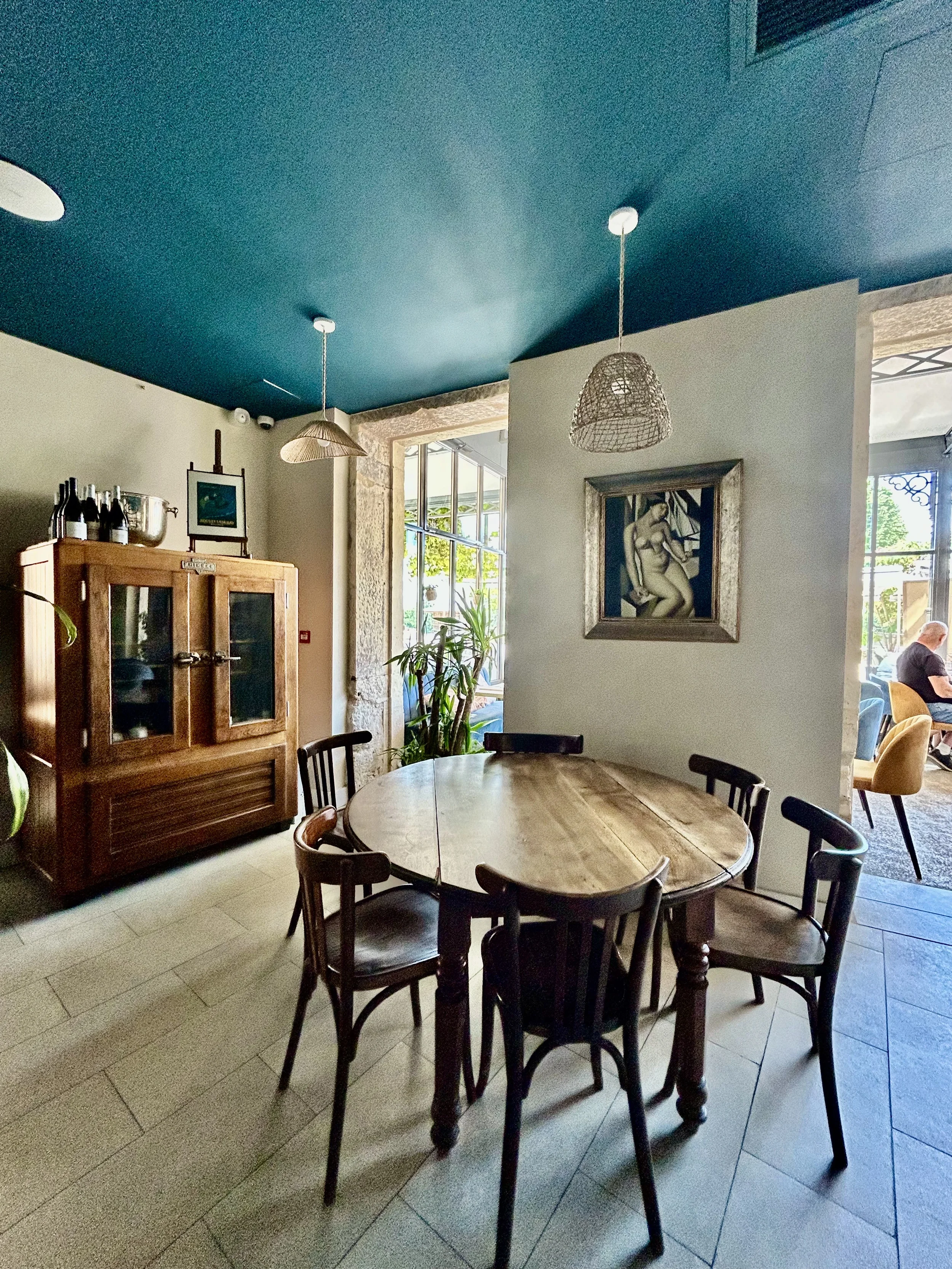 Intérieur d'un restaurant avec un tableau, des plantes, une armoire en bois, un plafond bleu et une table ronde en bois entourée de plusieurs chaises en bois.
